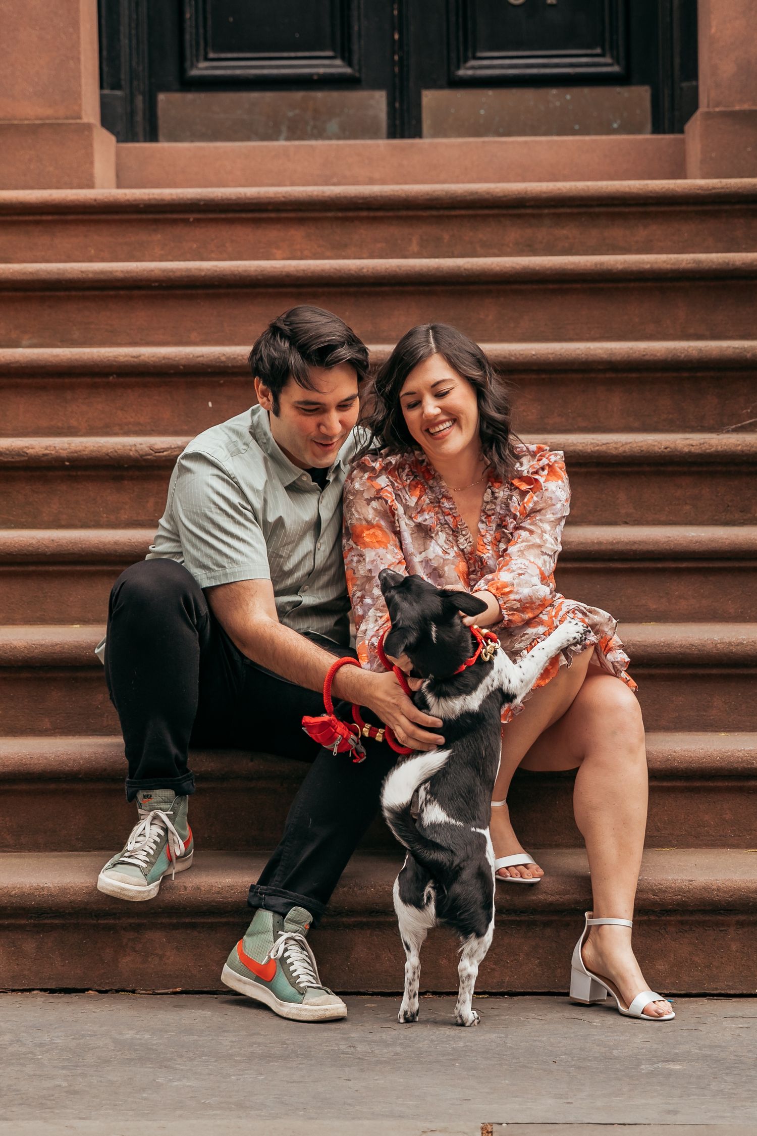 Smiling couple sitting on brownstone steps petting their small black and white dog together.