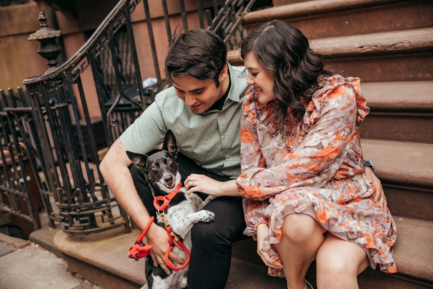 Couple on urban stoop playing with their small dog wearing a red bandana on steps.