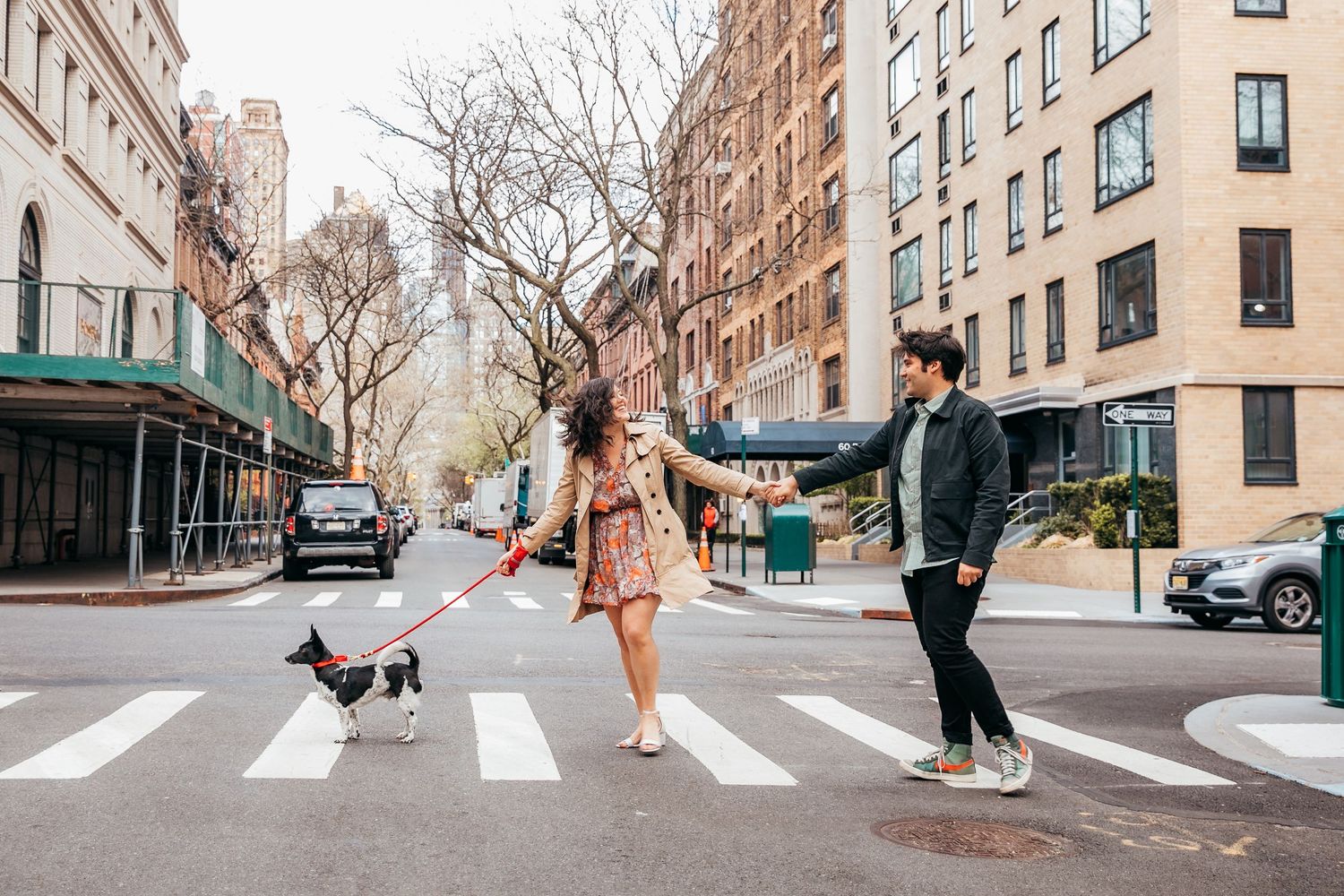 Couple crossing city street with small dog on red leash in New York neighborhood.