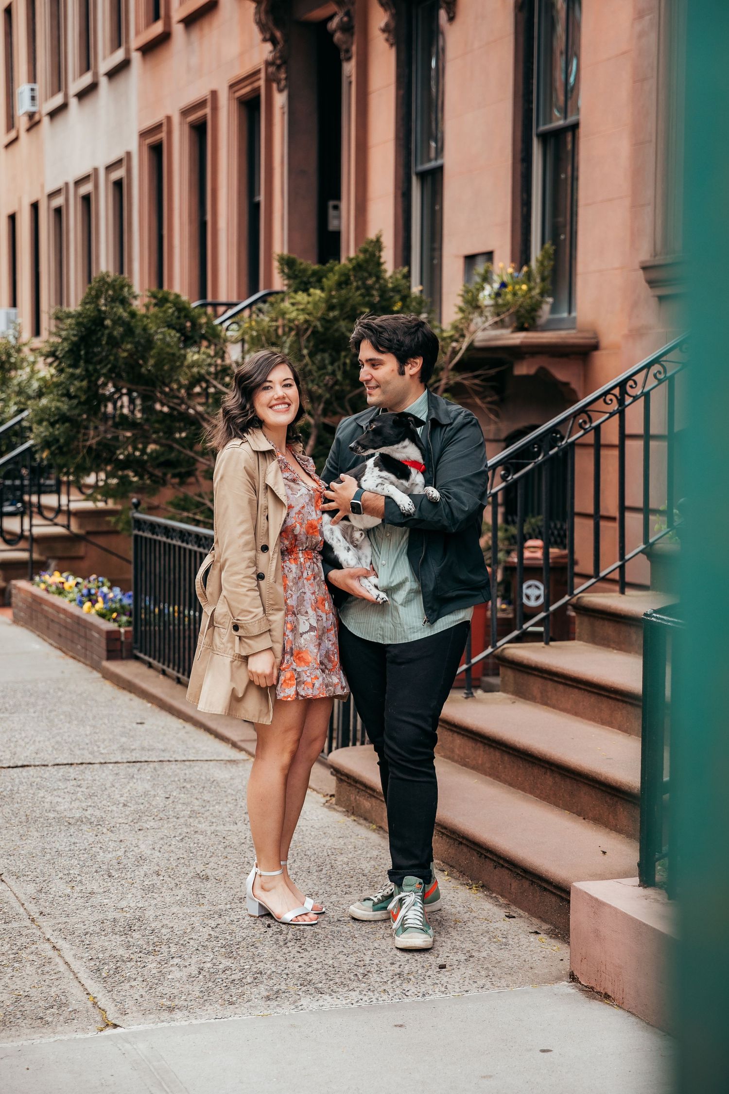 Smiling couple standing on New York City brownstone sidewalk with their small dog.