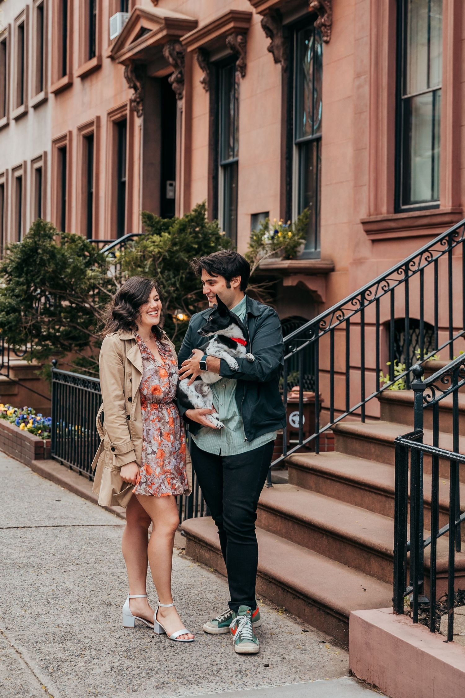 A couple smiles while holding a small dog on a sunny NYC brownstone sidewalk.