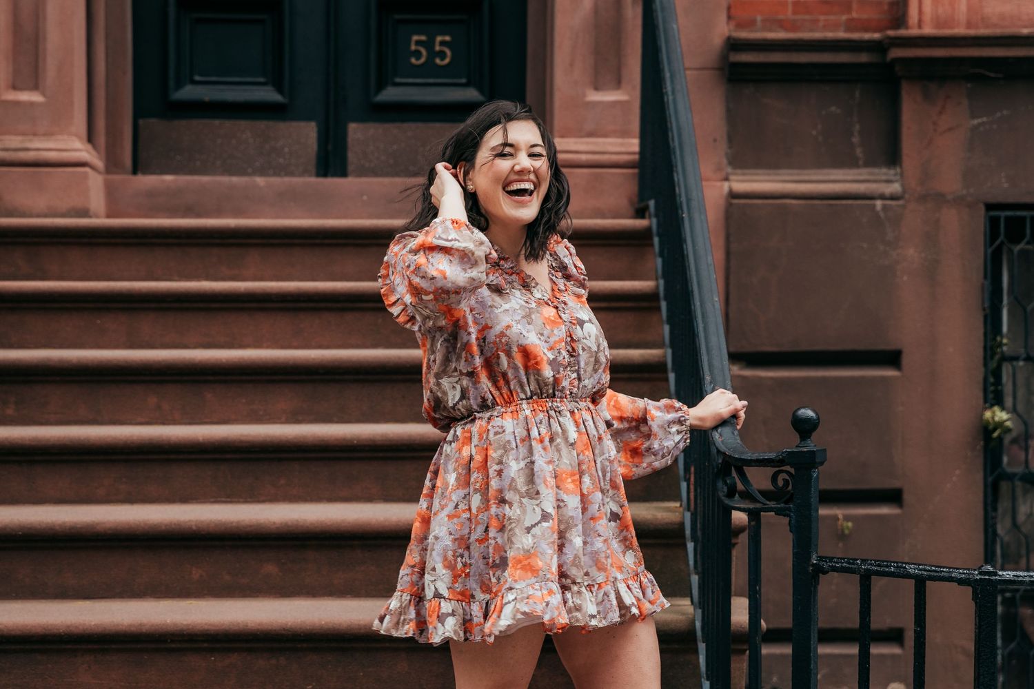 A joyful woman in a floral dress laughs on the steps of a NYC brownstone.