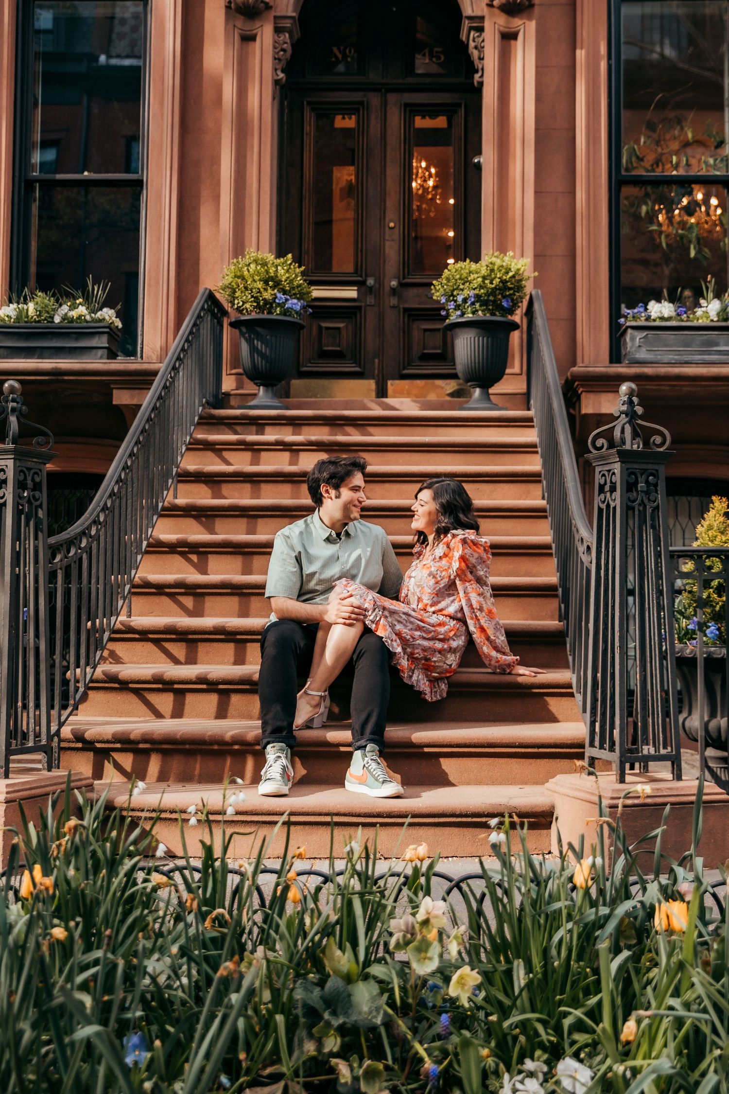 A romantic couple sits together on brownstone steps surrounded by spring flowers.