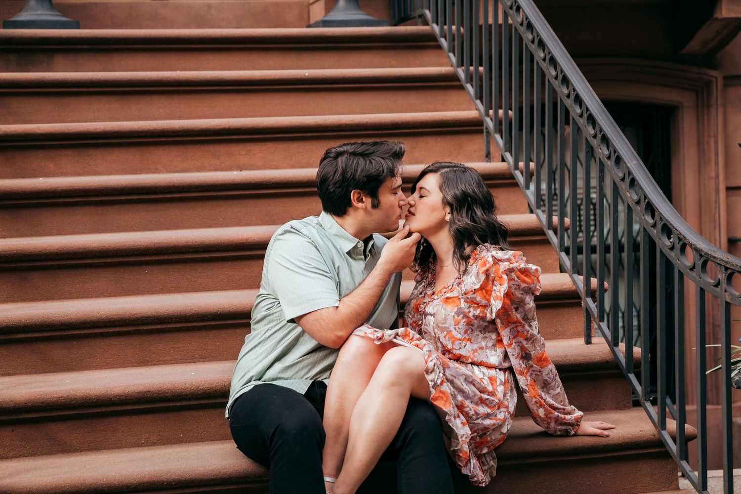 A couple shares a romantic kiss while seated on brownstone steps outdoors.