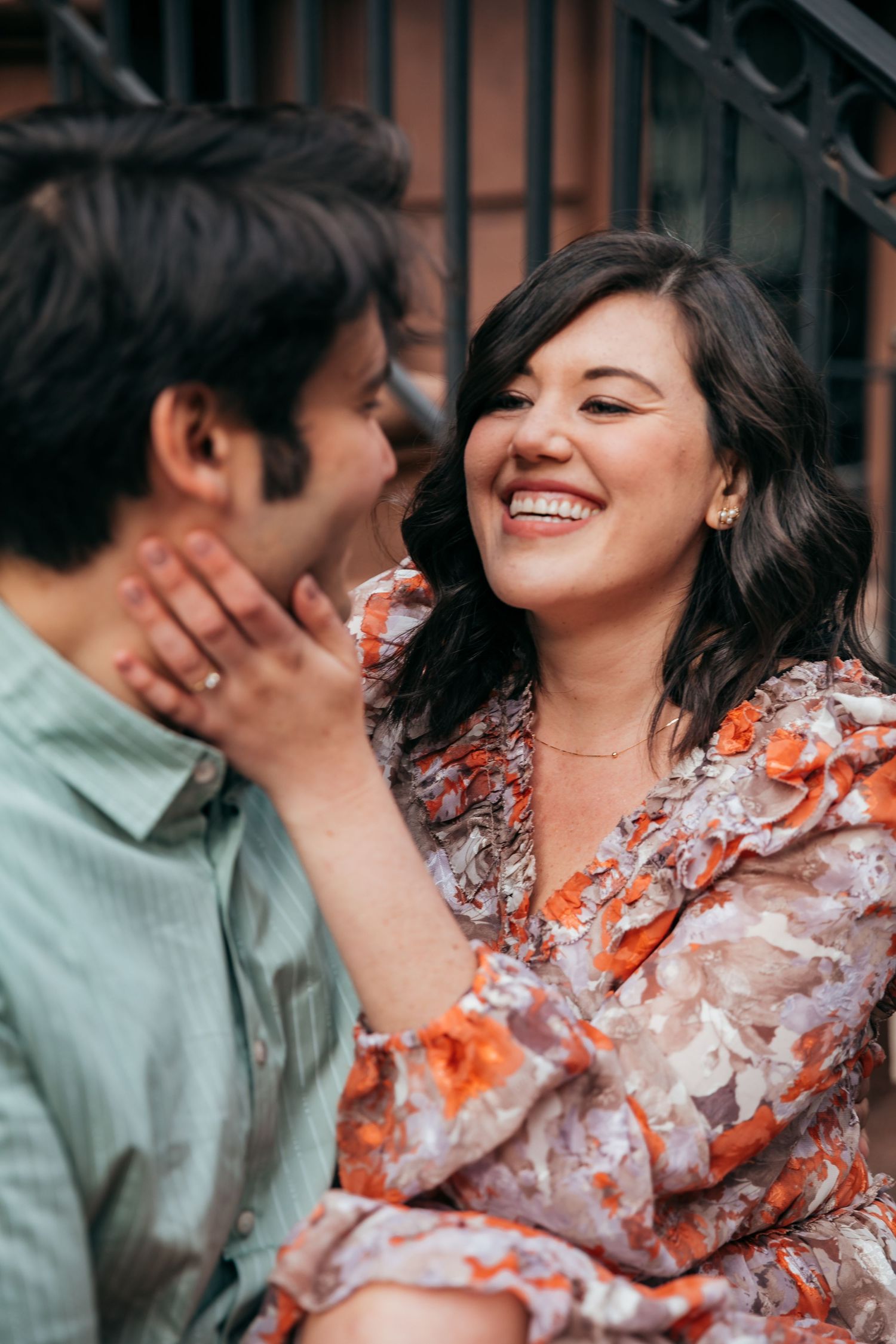 Woman in floral dress laughing joyfully while holding mans face during outdoor couples photo session.