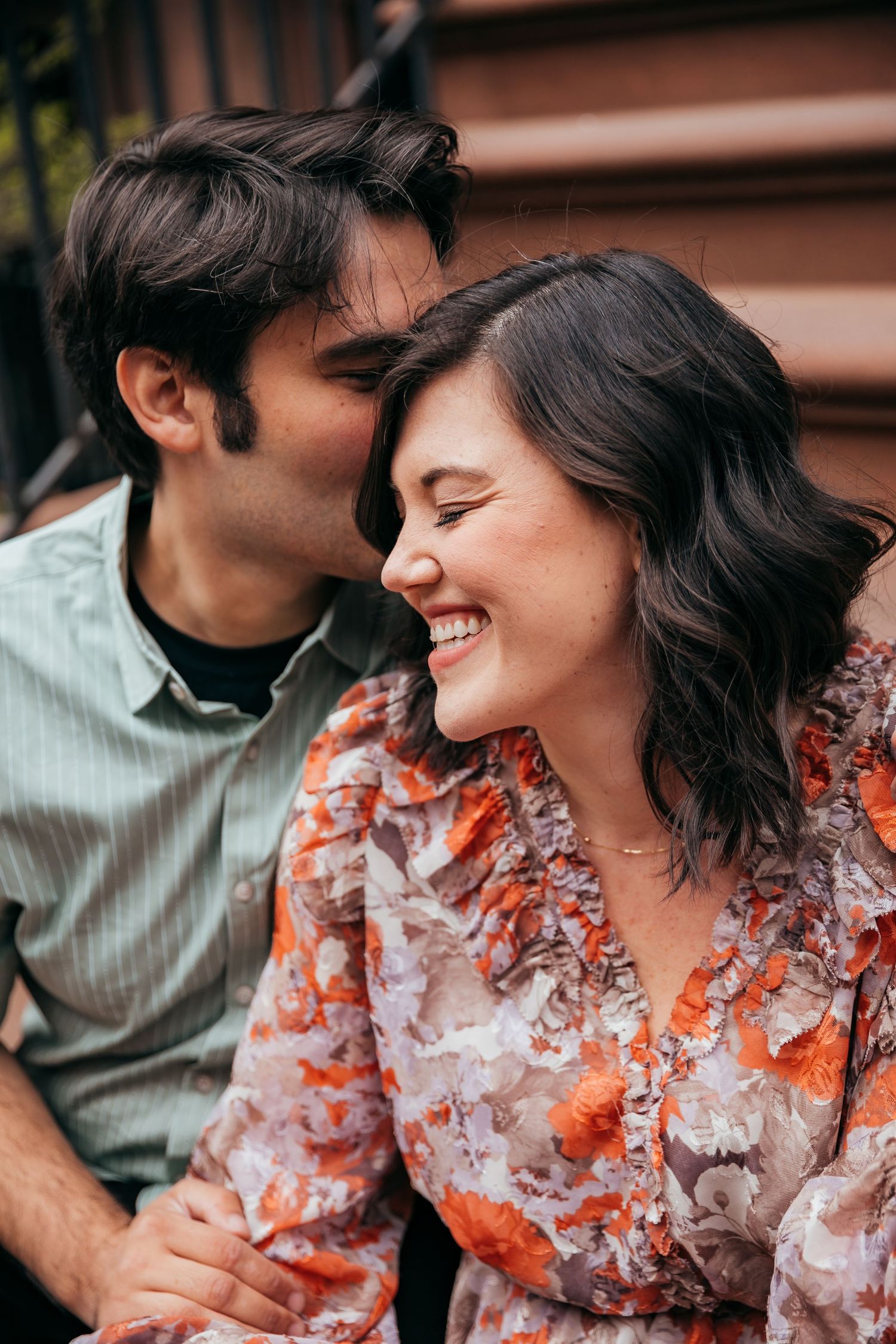 Man whispering to laughing woman in orange floral dress during romantic outdoor couples photoshoot.