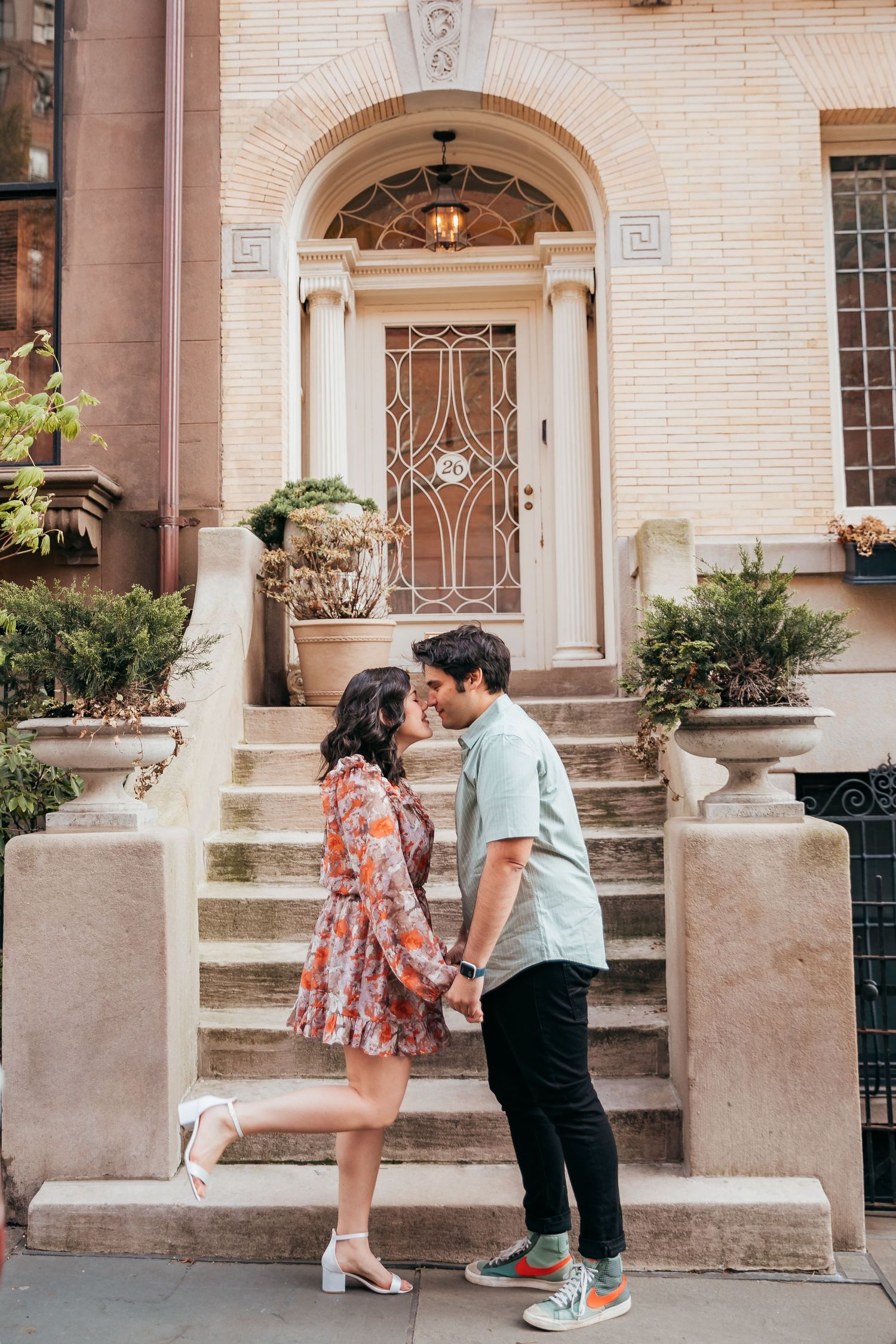 Couple leaning in for a kiss while holding hands in front of elegant townhouse entrance.