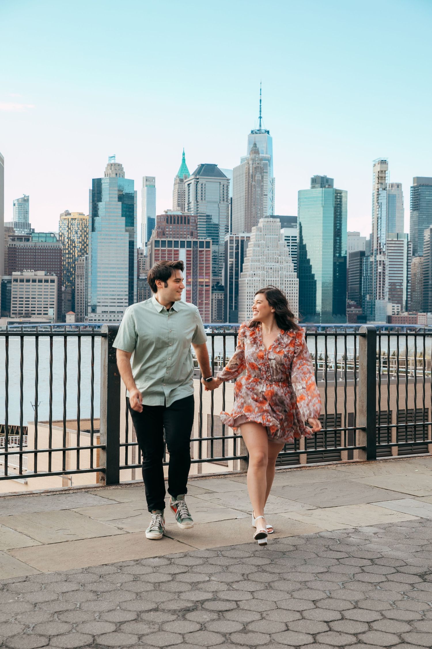 Smiling couple holding hands strolling along Brooklyn waterfront with Manhattan skyline.