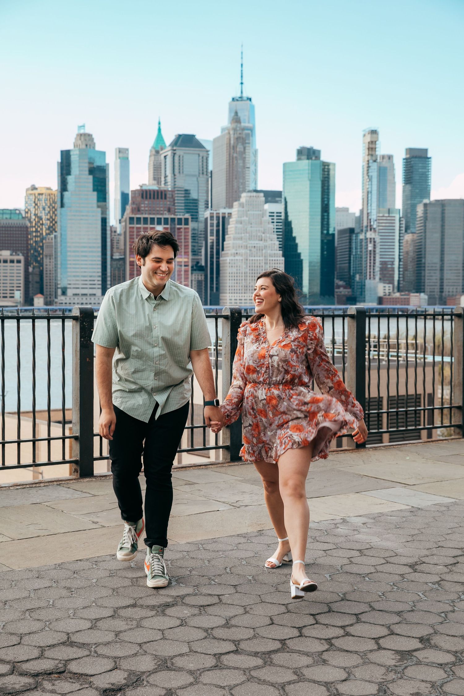 Laughing couple walking together on cobblestone path with New York City skyline behind.