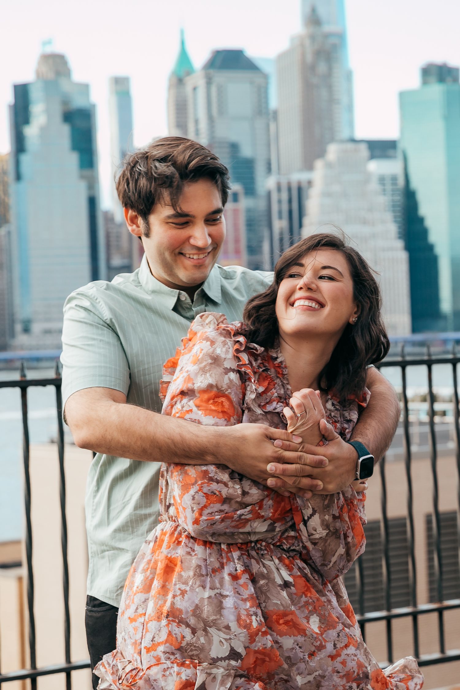 Man embracing woman in floral dress laughing together with NYC skyline behind them.