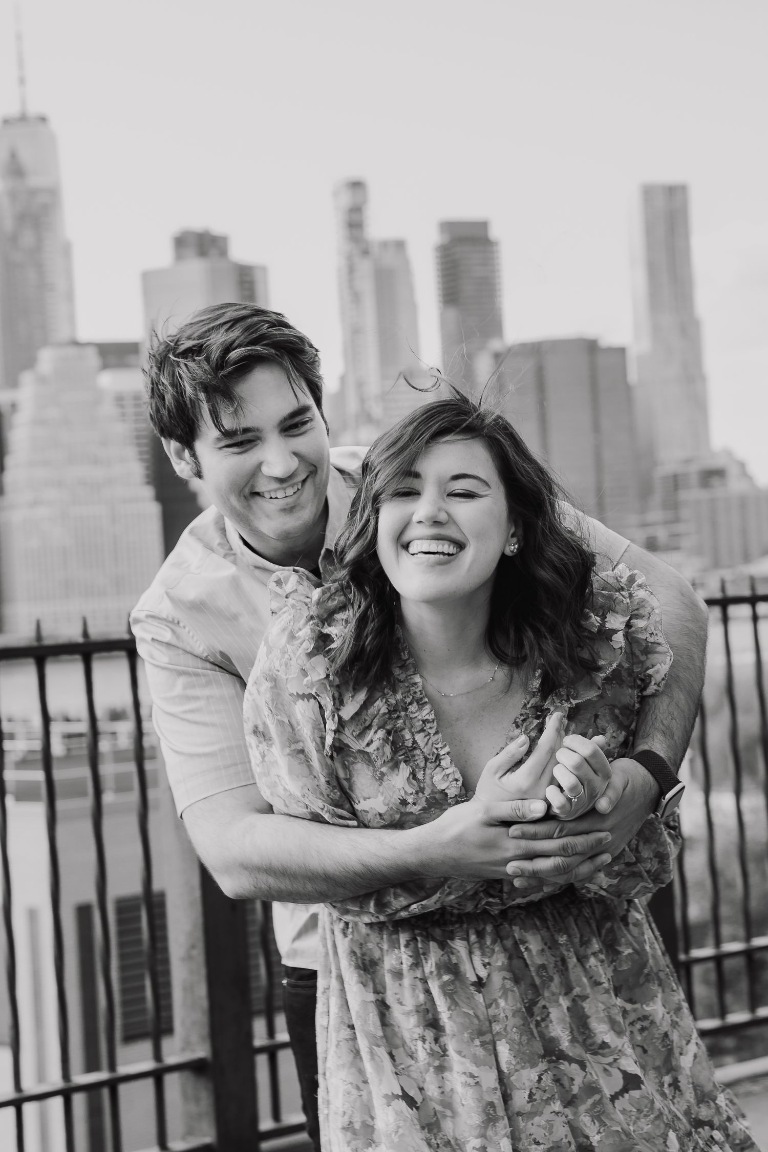 Black and white photo of smiling couple embracing with NYC skyline in background.