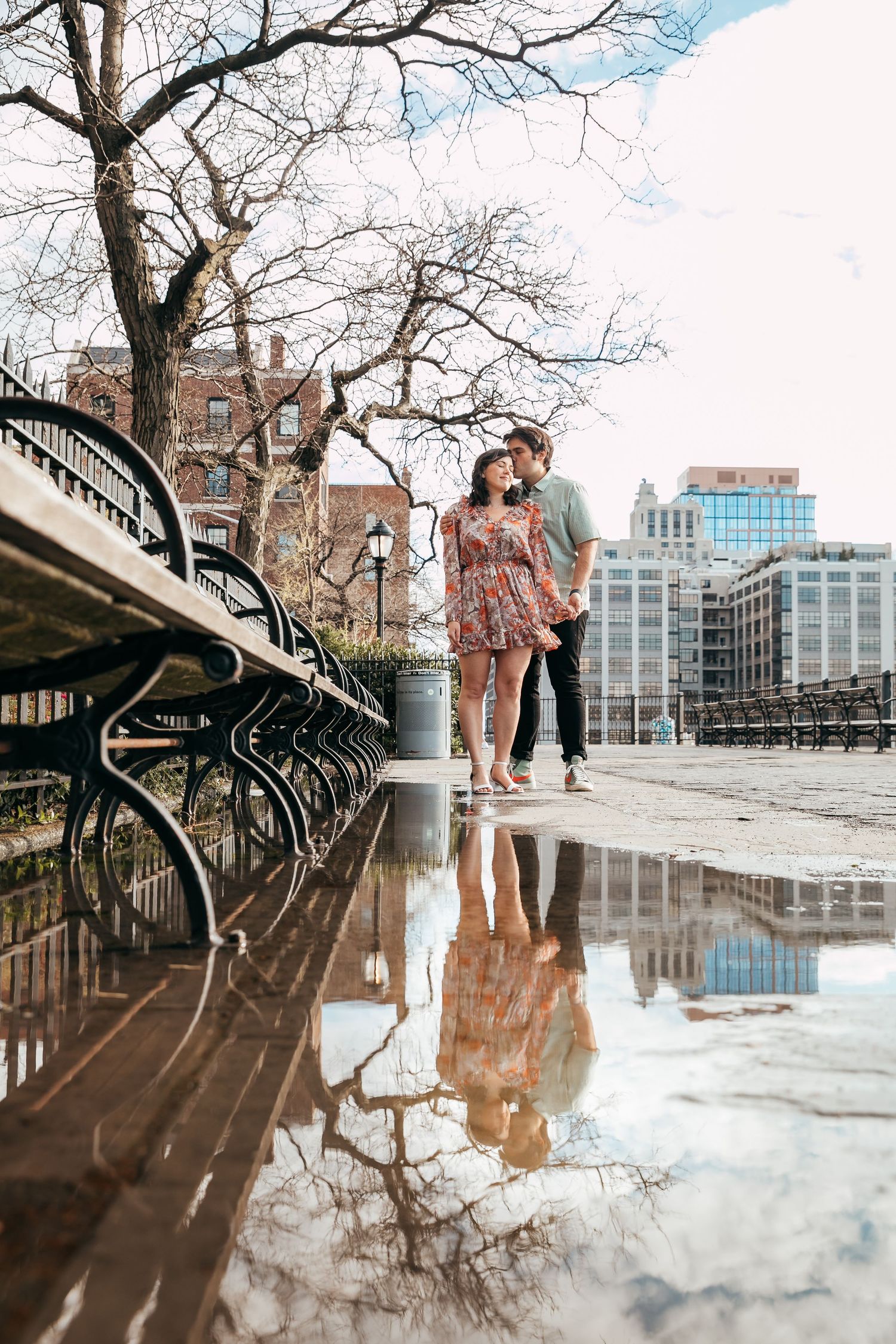 Couple sharing a kiss reflected in puddle near Brooklyn park benches and skyline.