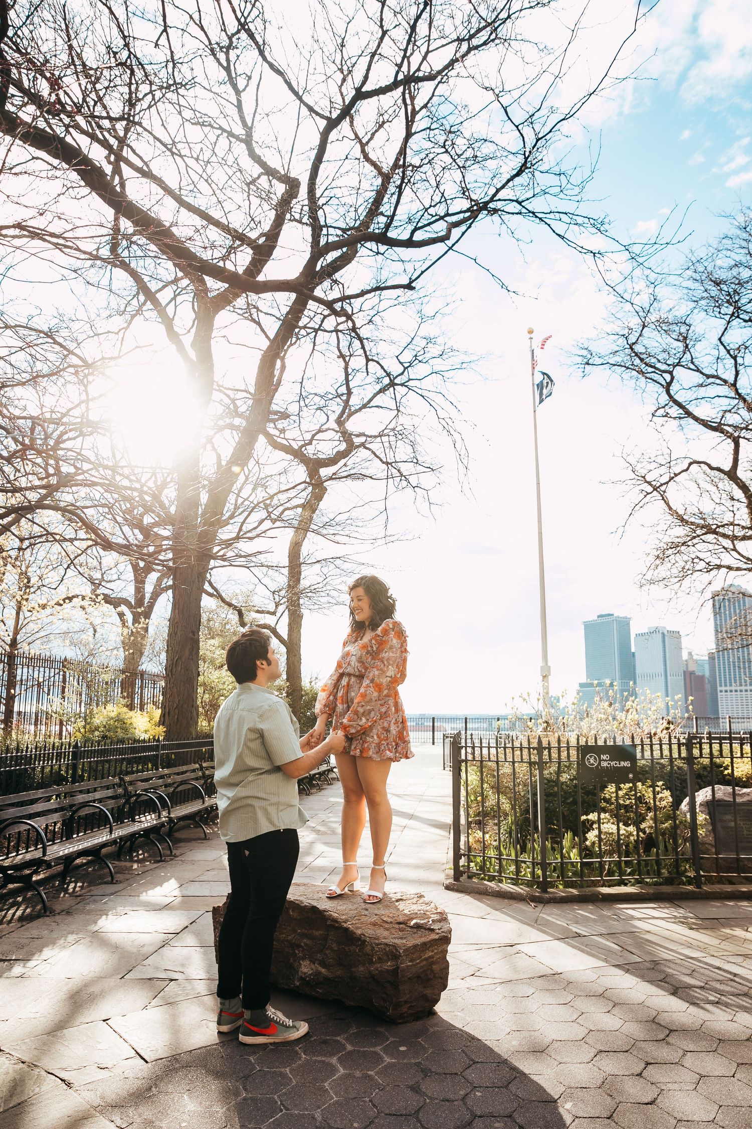 Couple sharing a romantic proposal moment in a sunlit park with city skyline views in the background.