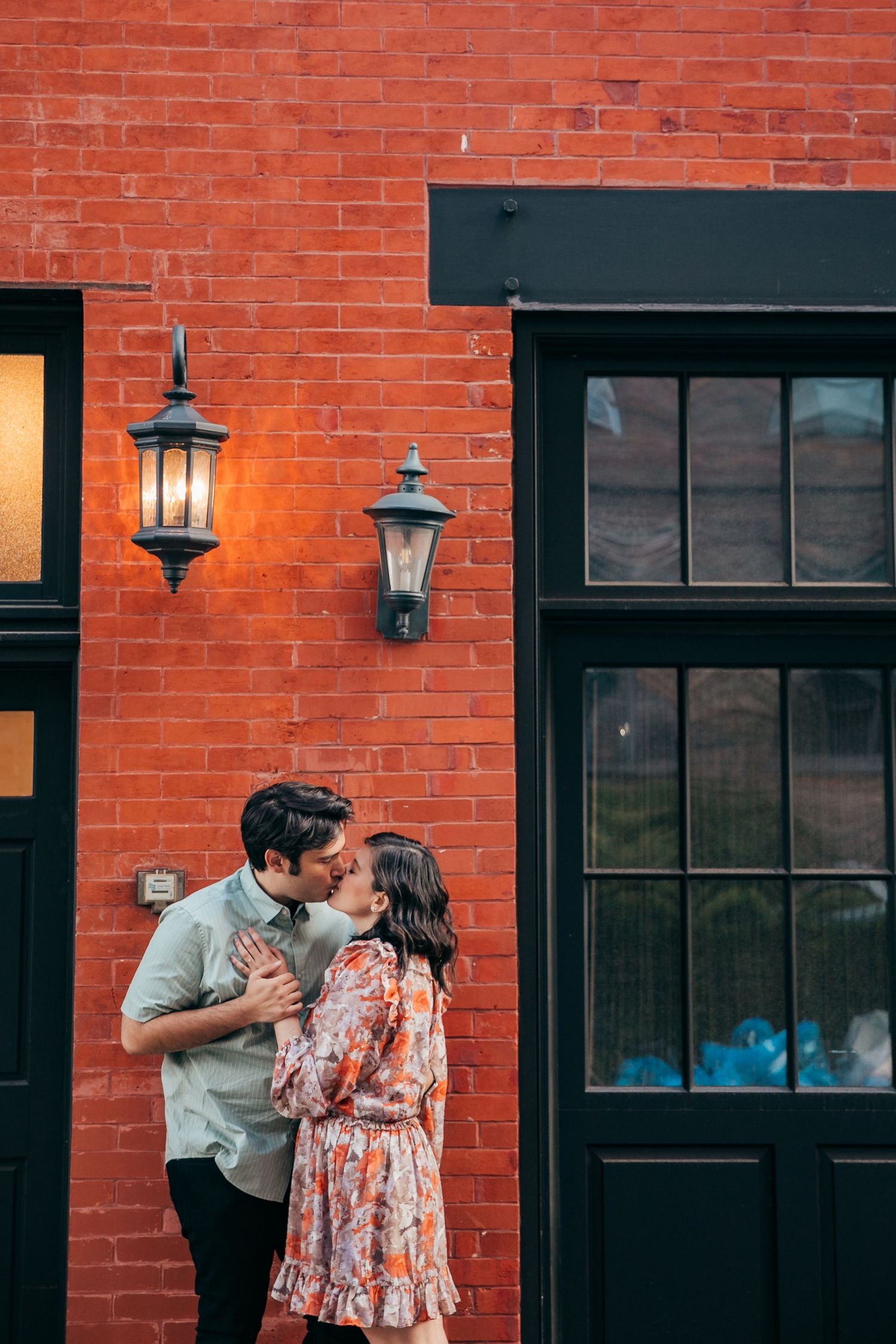 Couple sharing a tender kiss against a vibrant red brick wall with vintage lanterns in an urban setting.