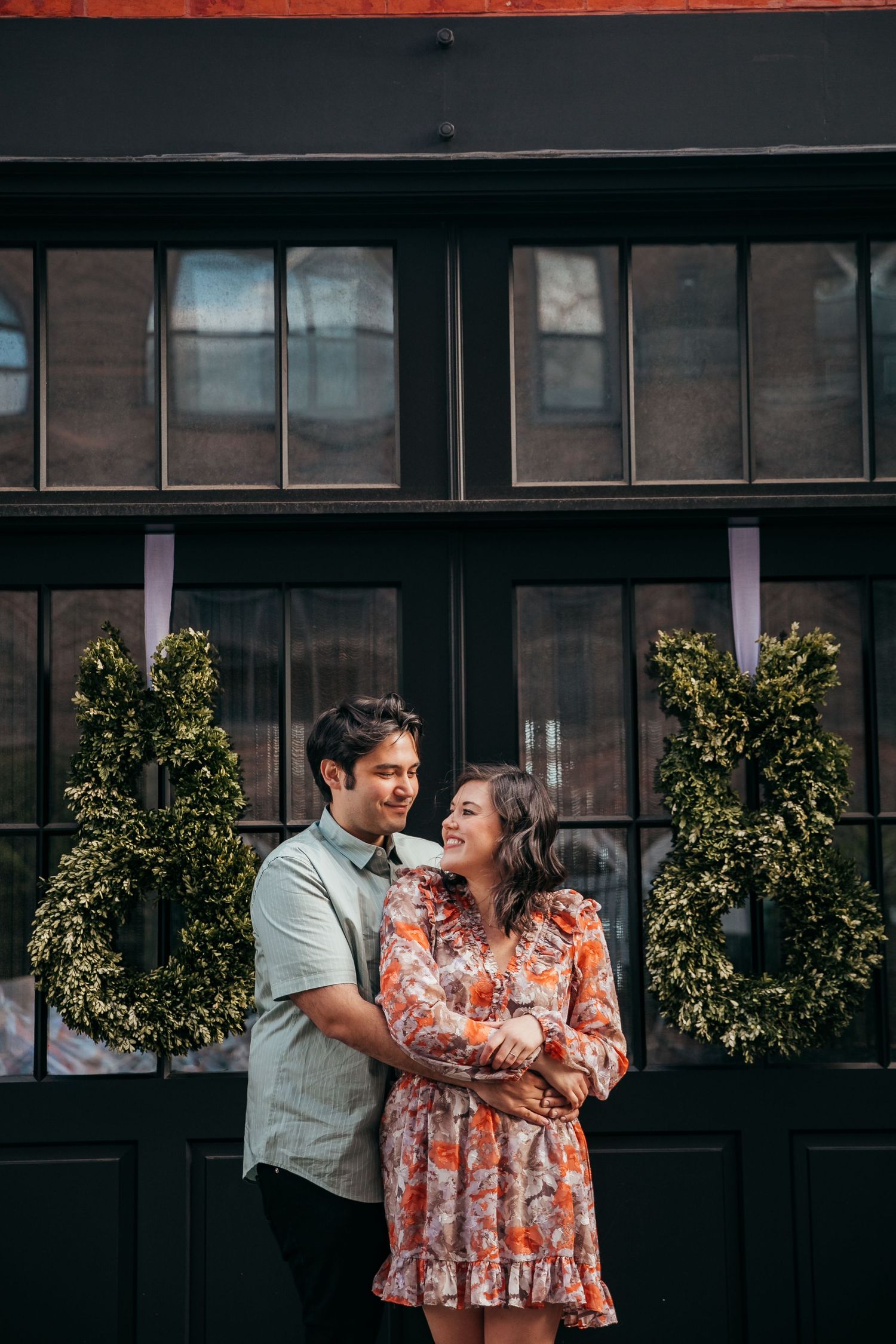 Happy couple embracing and laughing together in front of a dark storefront adorned with green topiaries.