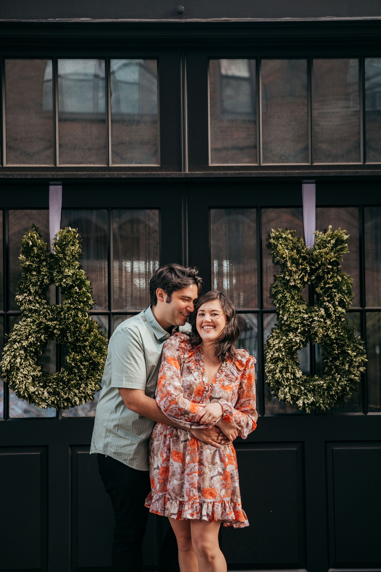 Smiling couple cuddling closely in front of elegant black windows decorated with lush boxwood wreaths.