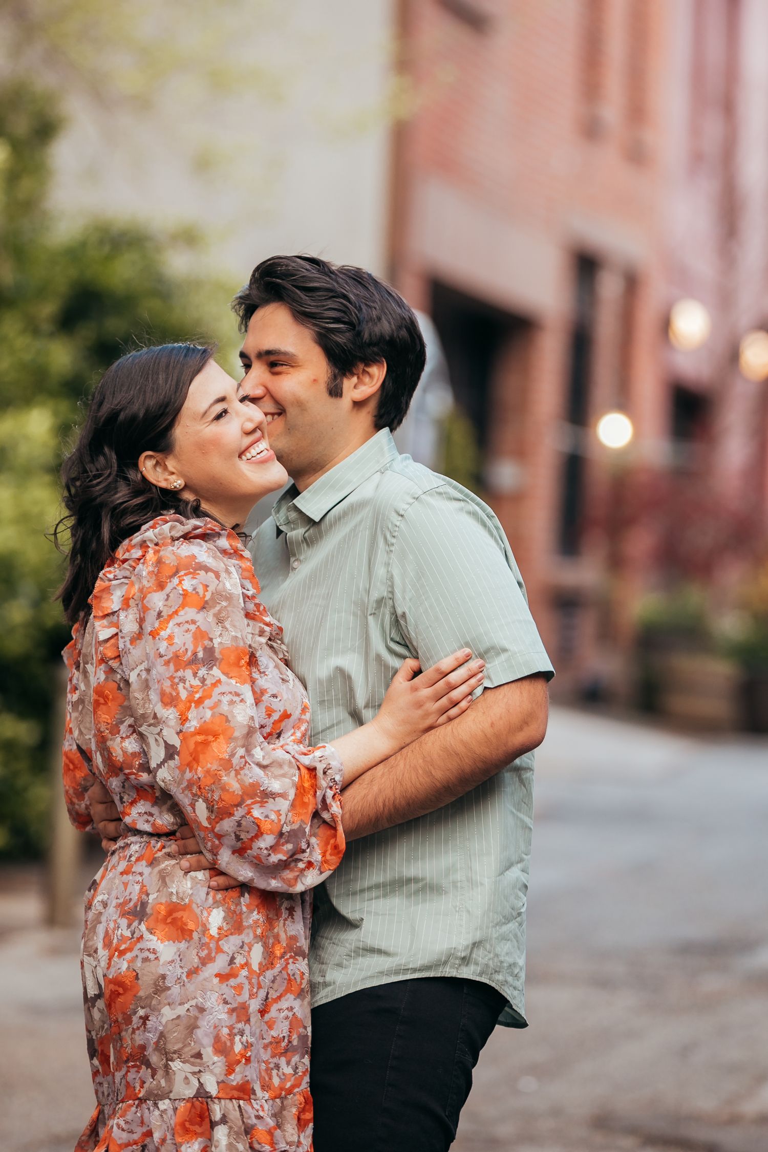 Laughing couple holding each other close on a picturesque urban alley lined with warm string lights.