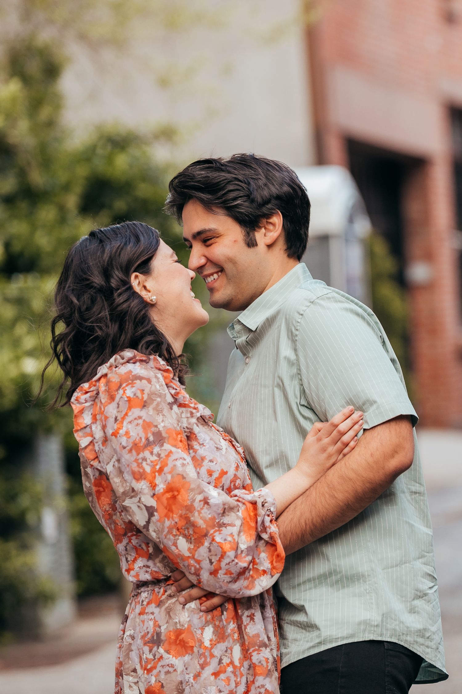 Smiling couple embracing and gazing at each other outdoors near greenery and brick building.