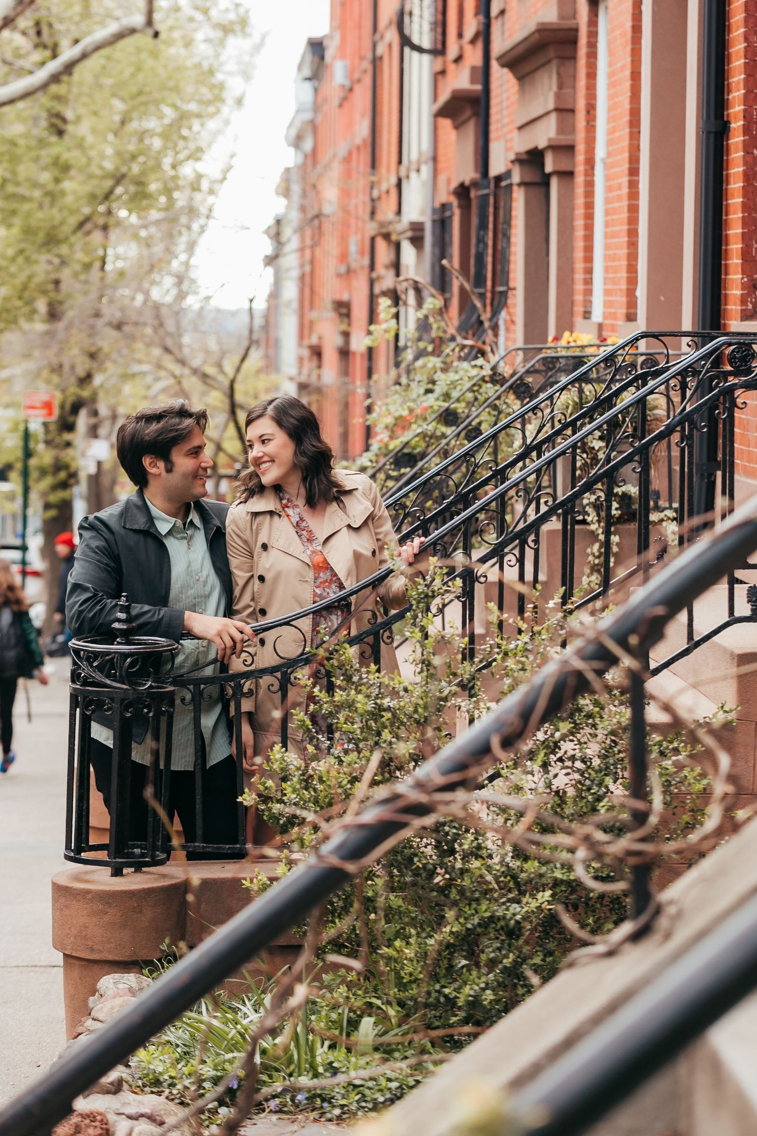 Happy couple holding hands on a tree-lined urban sidewalk with classic brownstone buildings.