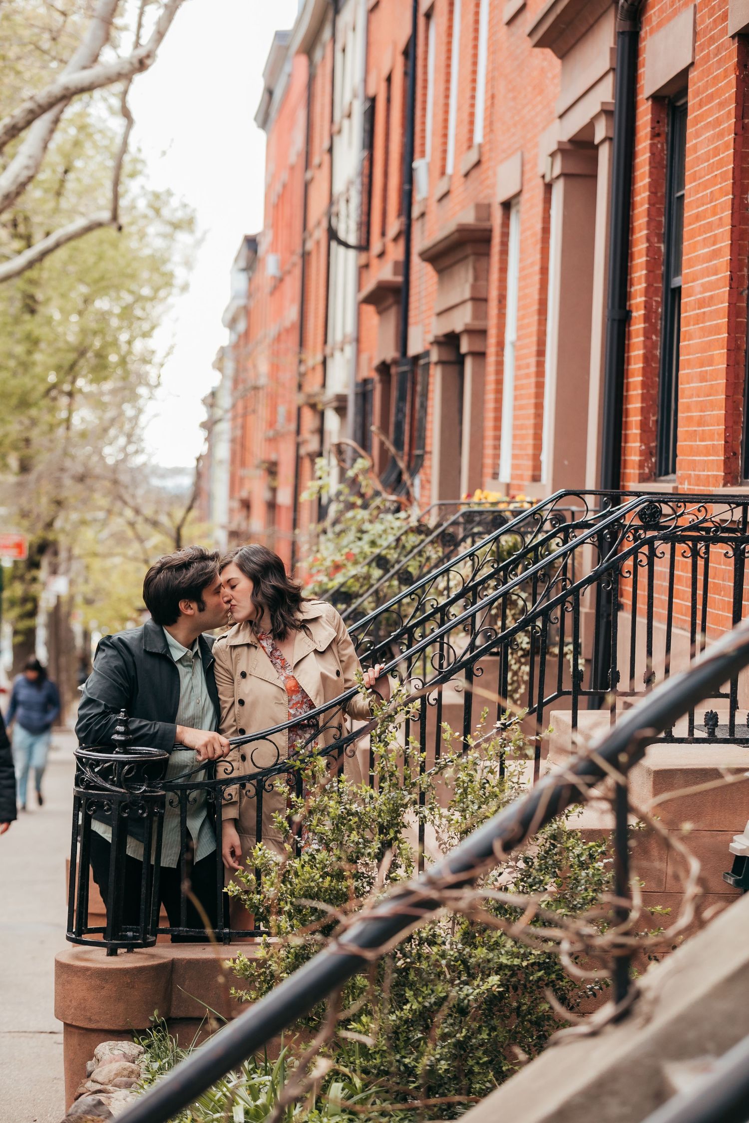 Couple kissing near iron railings on a classic urban brownstone-lined city sidewalk.