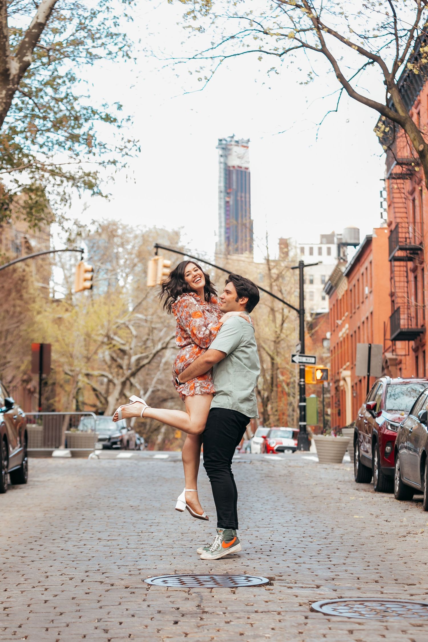 Joyful man lifting smiling woman on a cobblestone city street with urban skyline backdrop.