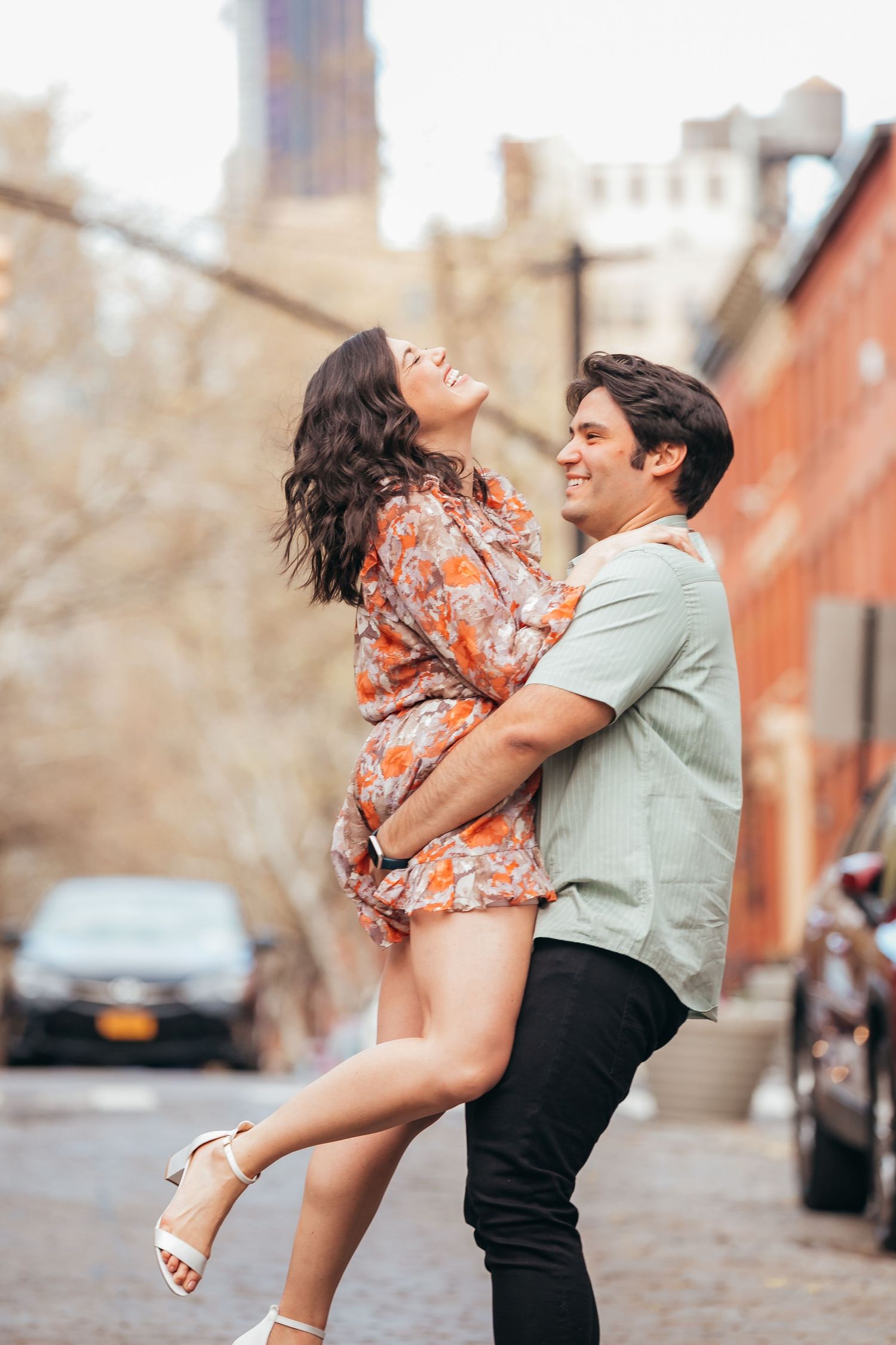 Man lifting laughing woman in floral dress on a cobblestone New York City street.
