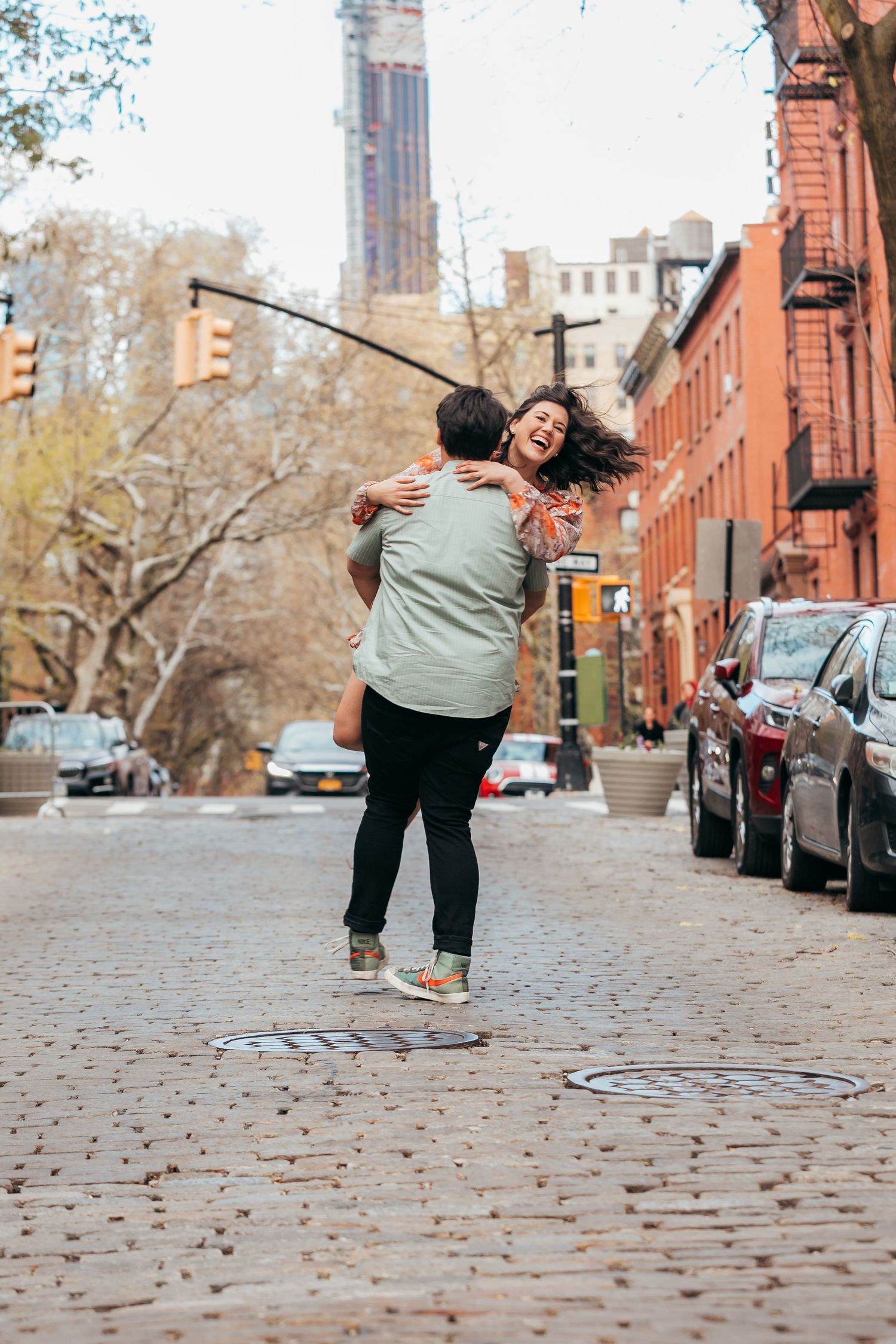 Man spinning laughing woman on a cobblestone street lined with brownstones and parked cars.