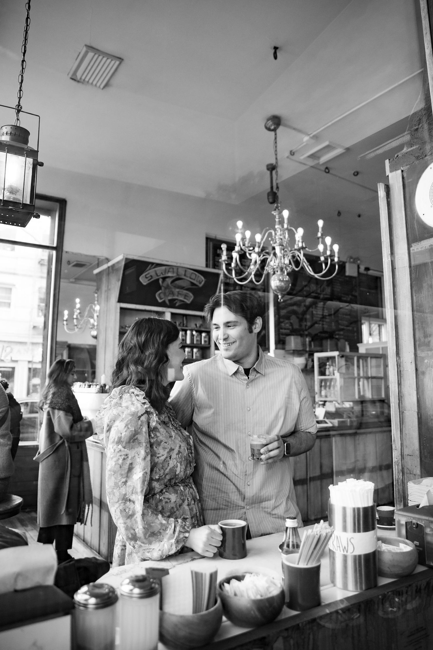 Black and white photo of a couple smiling at each other inside Swallow Cafe with a chandelier above.