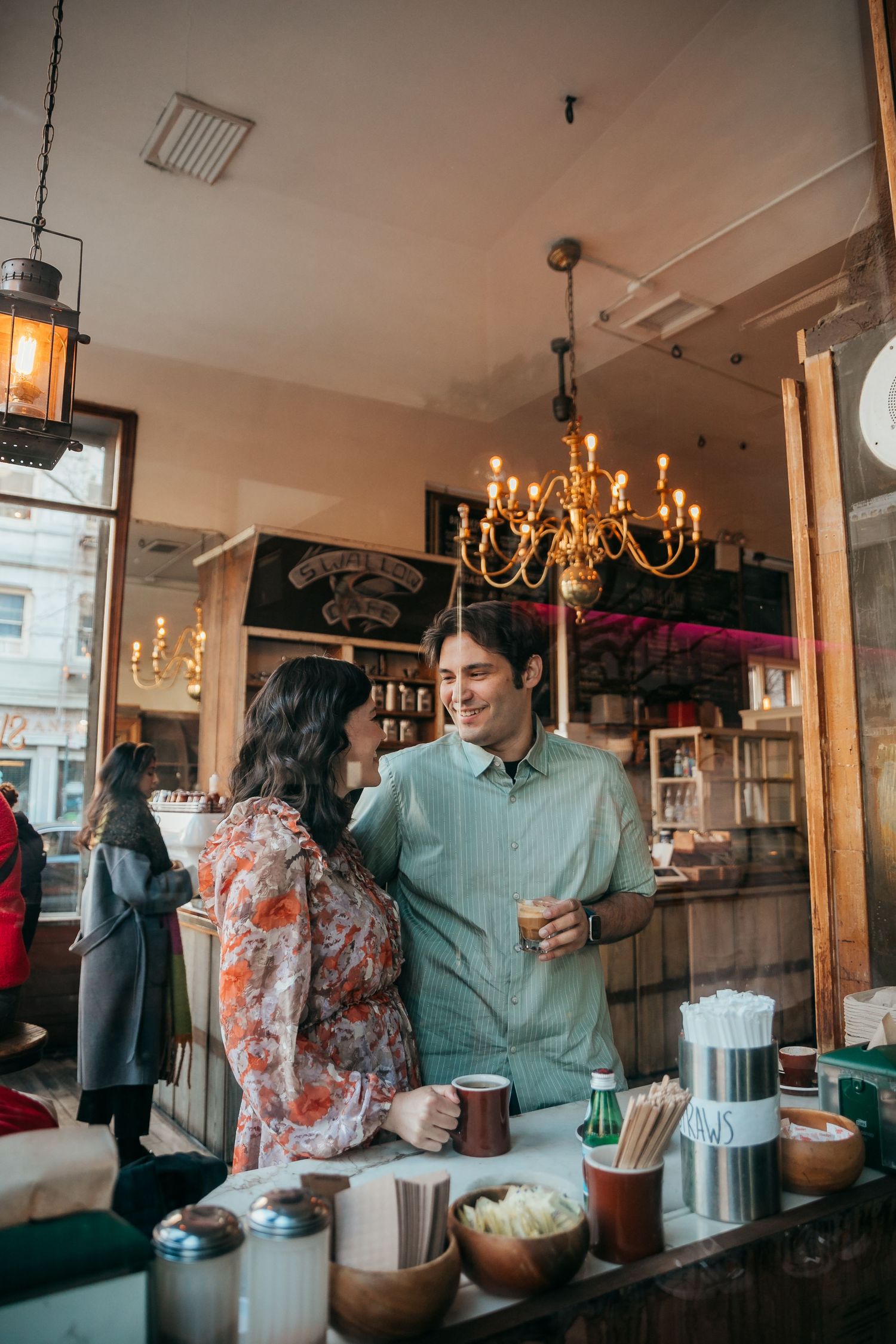 A couple shares a warm moment over coffee inside a cozy cafe with a golden chandelier overhead.