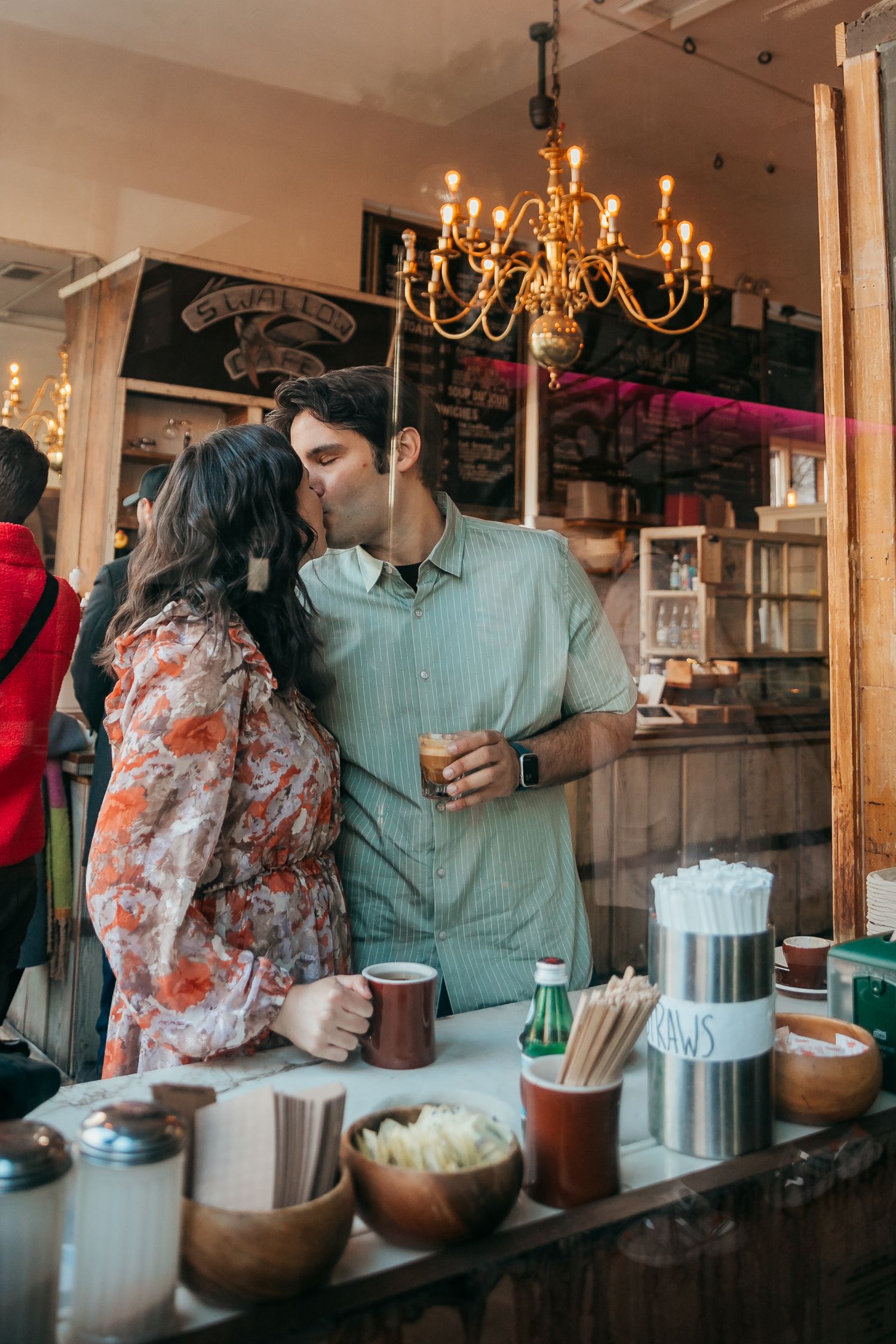 A couple shares a romantic kiss inside a cafe while holding their drinks at the counter.