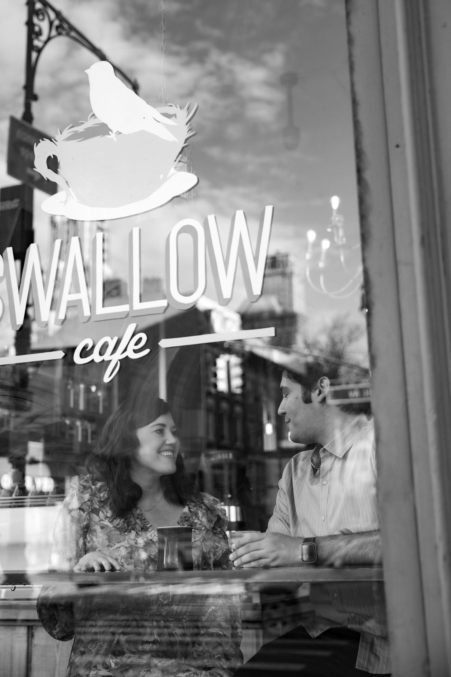 Black and white photo of a couple chatting inside Swallow Cafe, seen through the window.