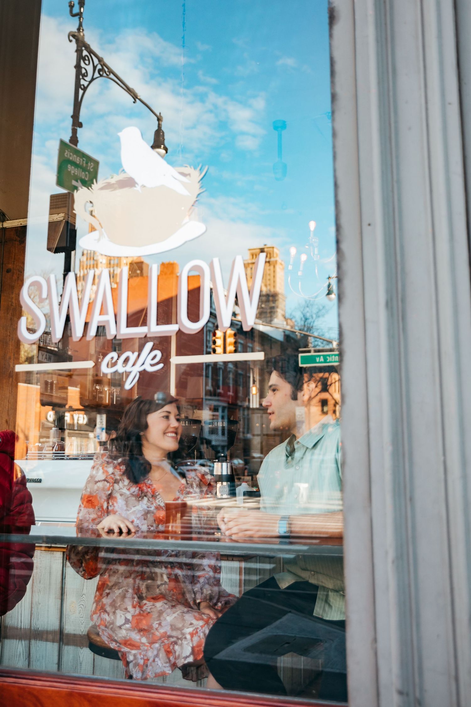 A couple enjoys conversation inside Swallow Cafe, photographed through the window on a sunny day.