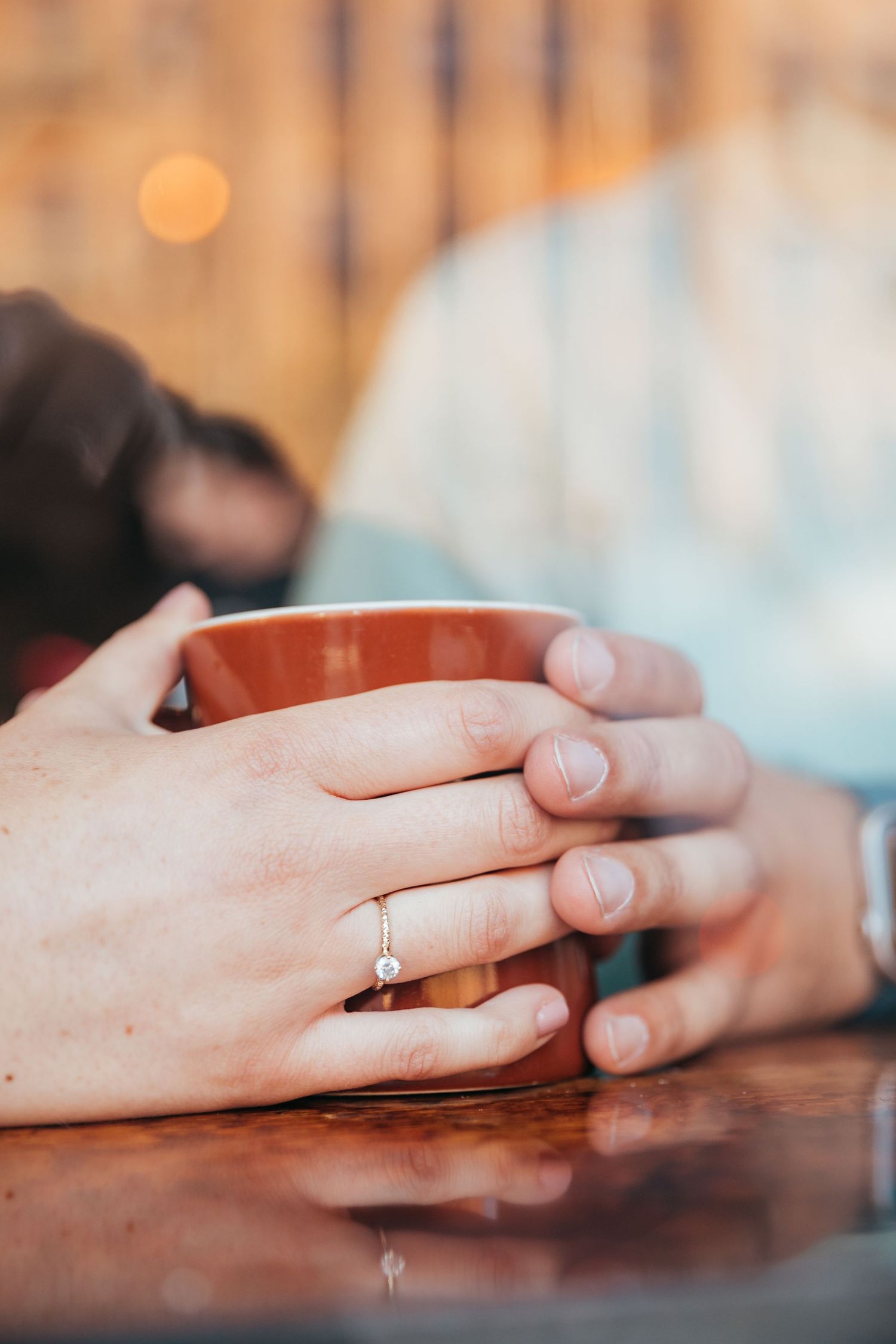 Close-up of two hands holding a warm mug, with an engagement ring visible on one hand.