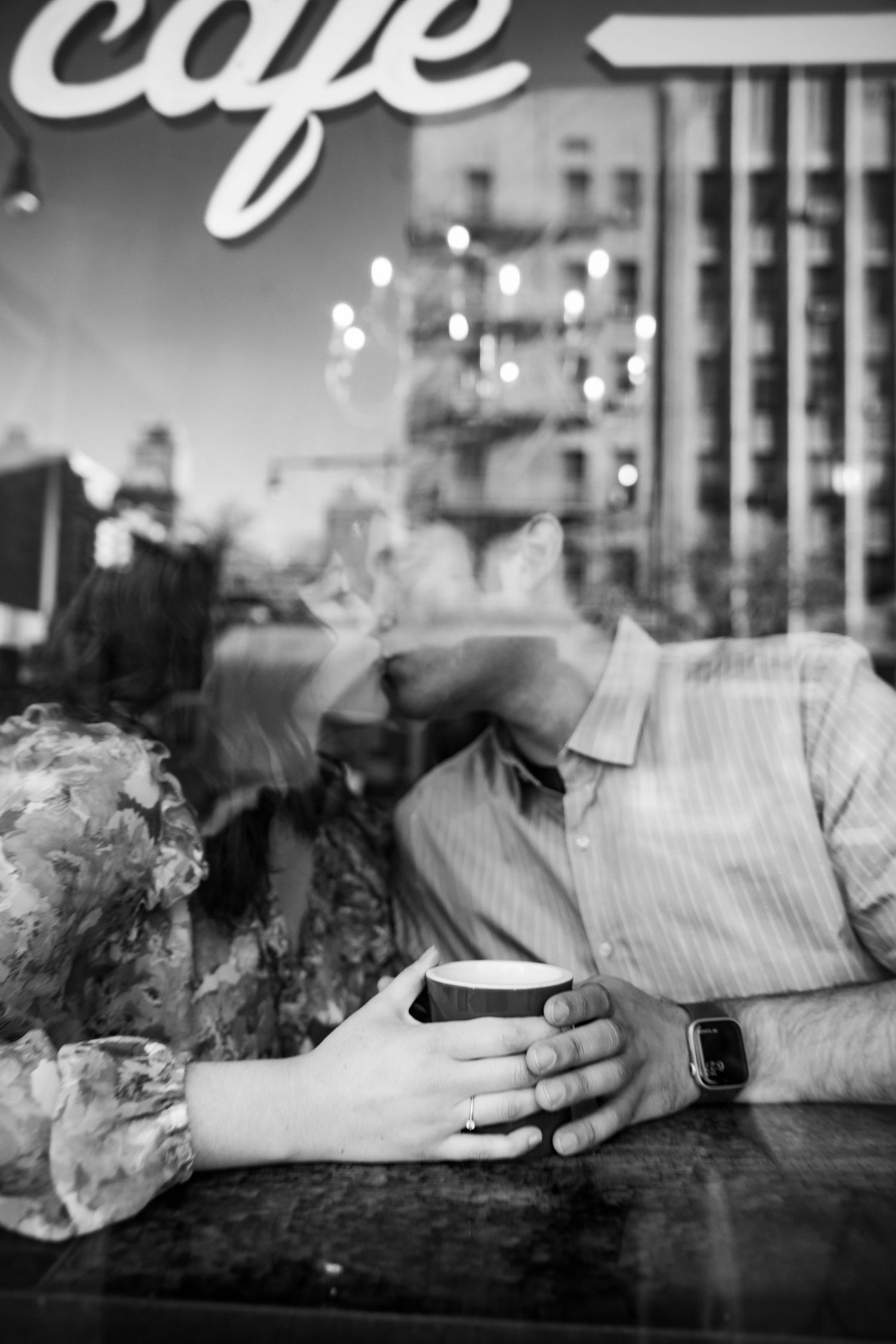 Black and white window reflection shot of a couple holding coffee cups inside a cafe.