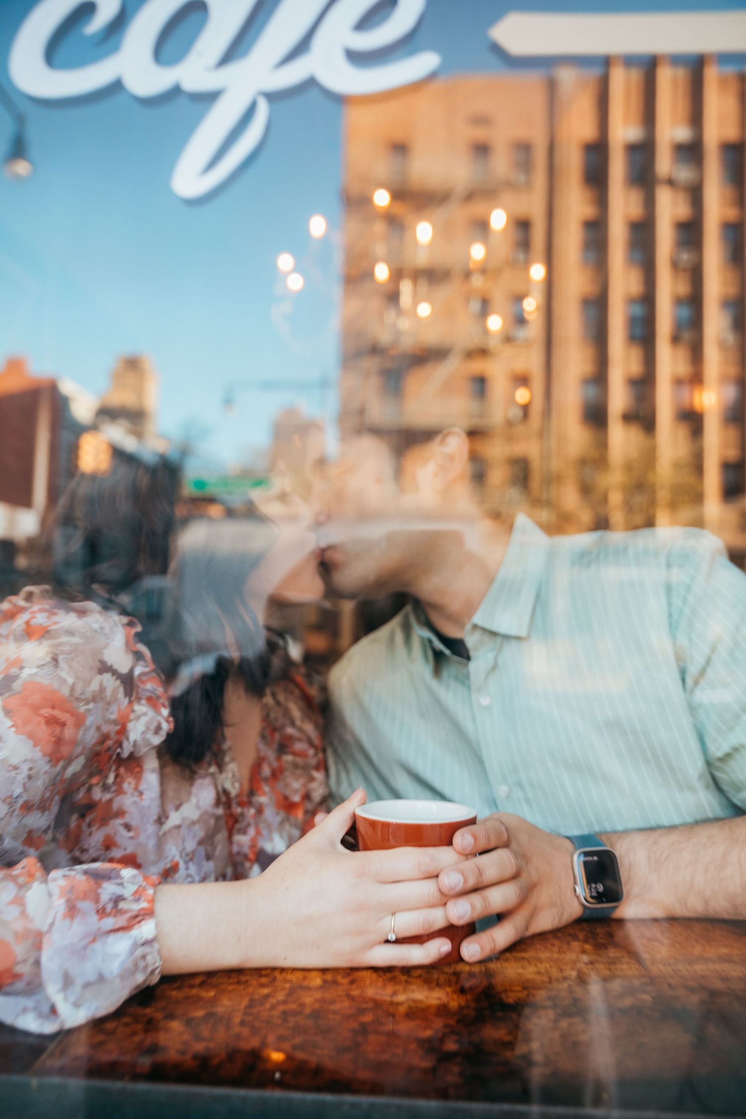 A couple holds hands over a steaming coffee cup, photographed through a cafe window at dusk.