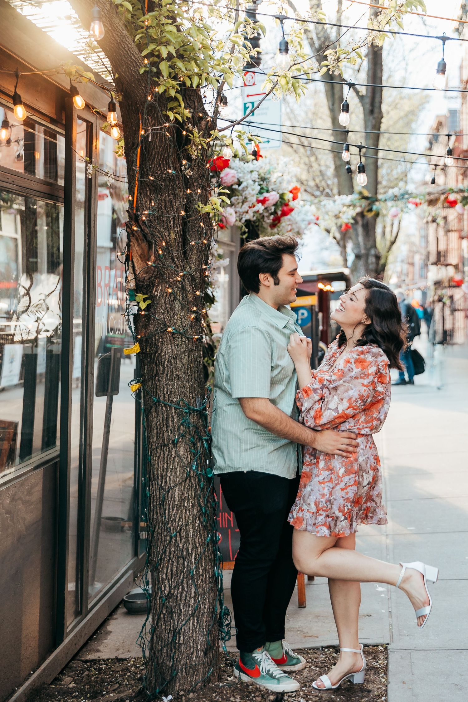 A joyful couple embraces beside a flower-decorated tree on a charming city sidewalk in spring.
