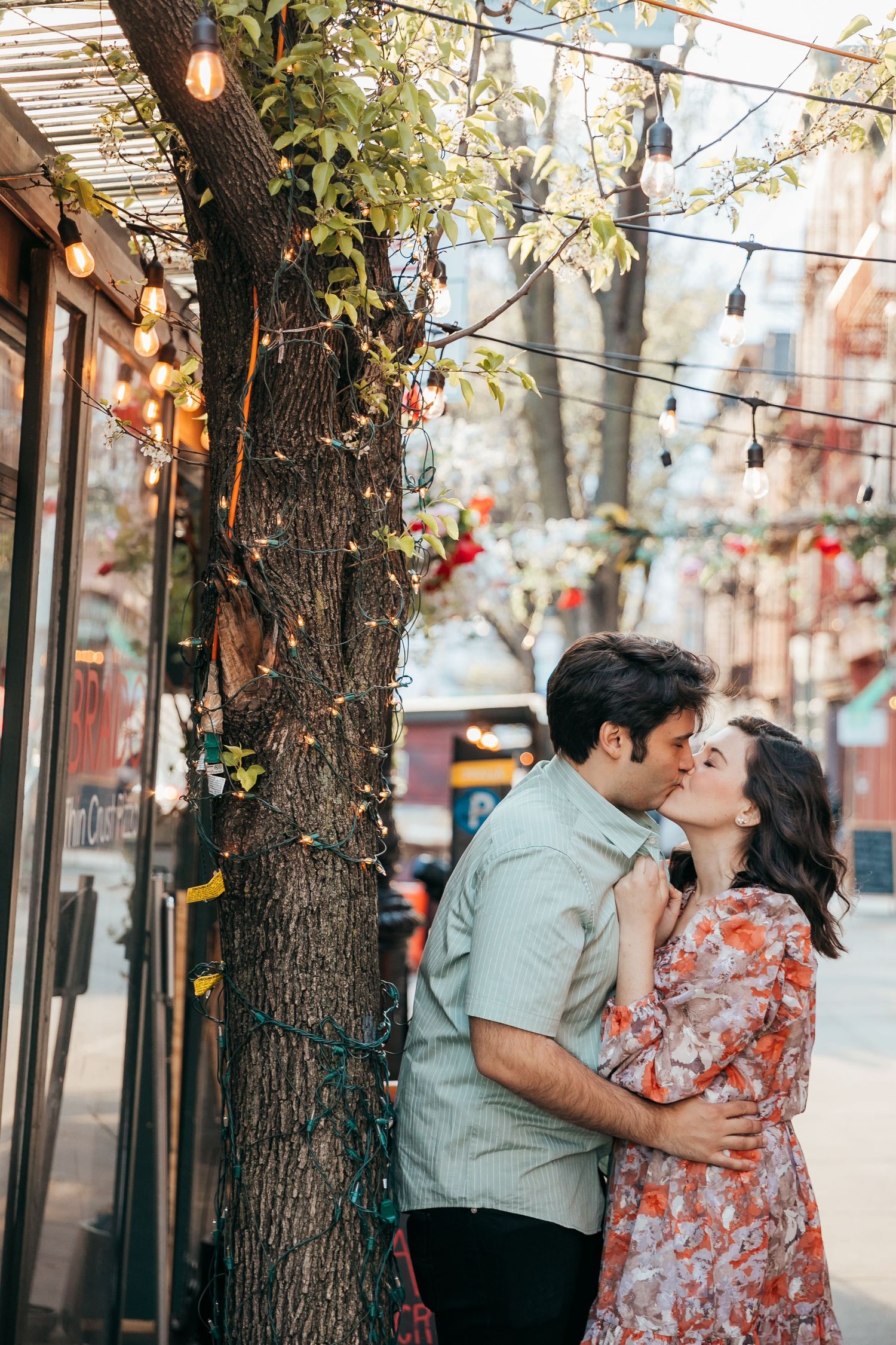 A couple shares a tender kiss beside a tree with string lights on a city street.