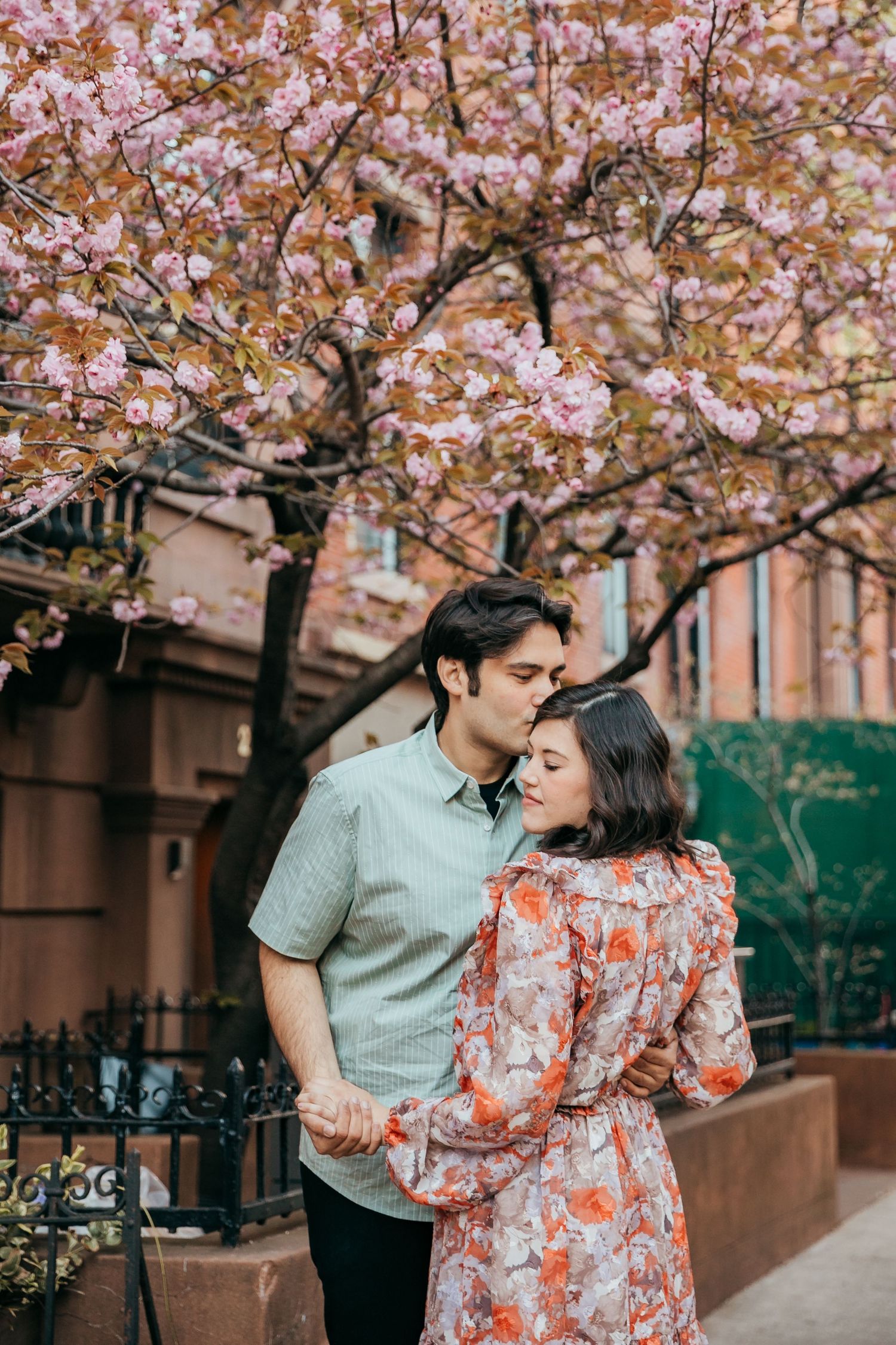 Couple embracing under pink cherry blossom tree on urban street, man kissing woman's forehead in spring.
