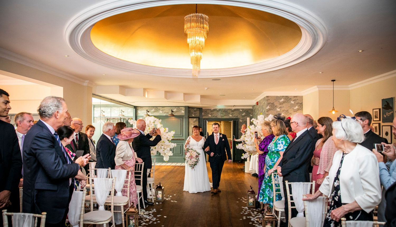 Panoramic view of wedding reception hall with guests and decorative lighting.