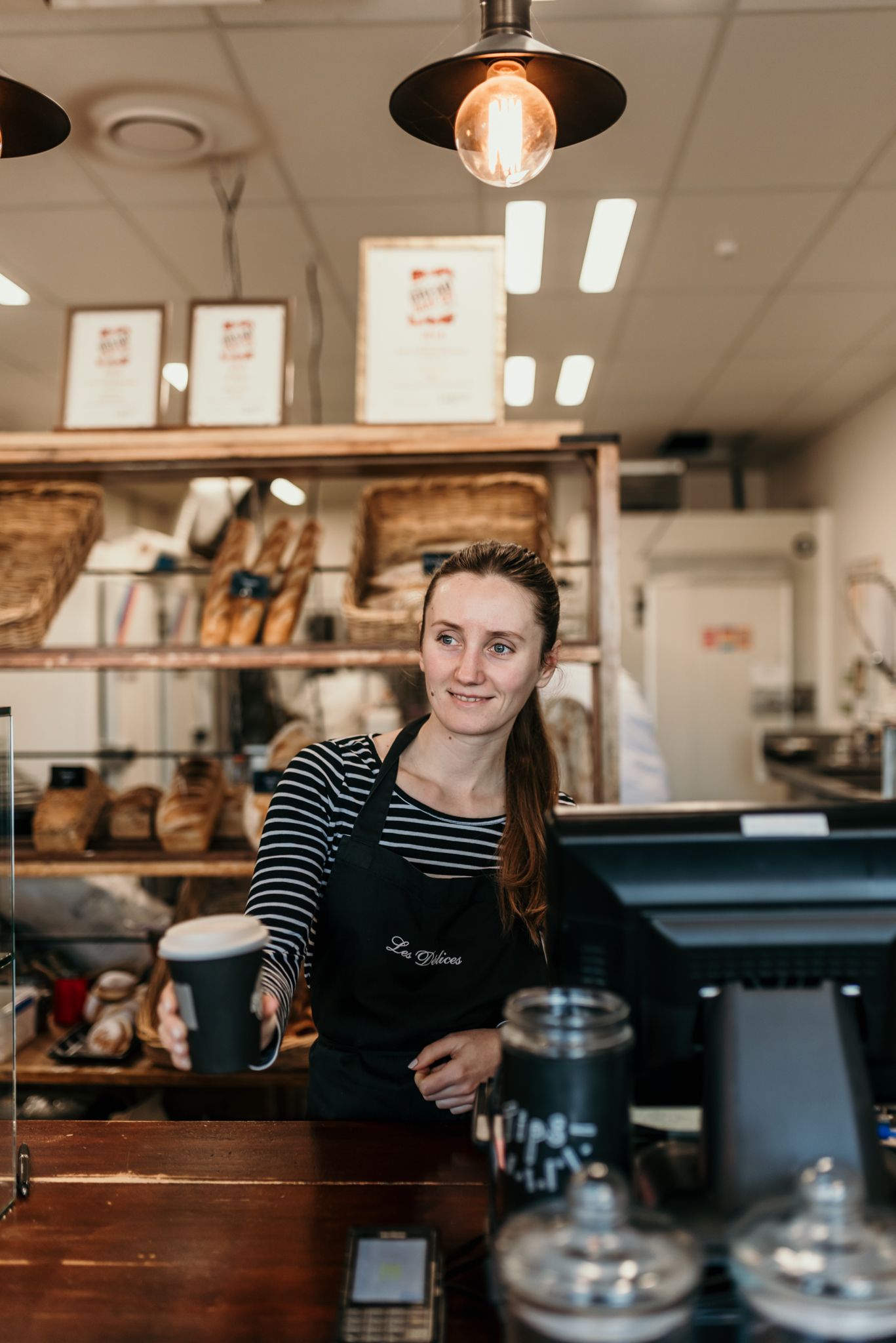 Staff member in striped shirt and black apron working behind counter at bakery.
