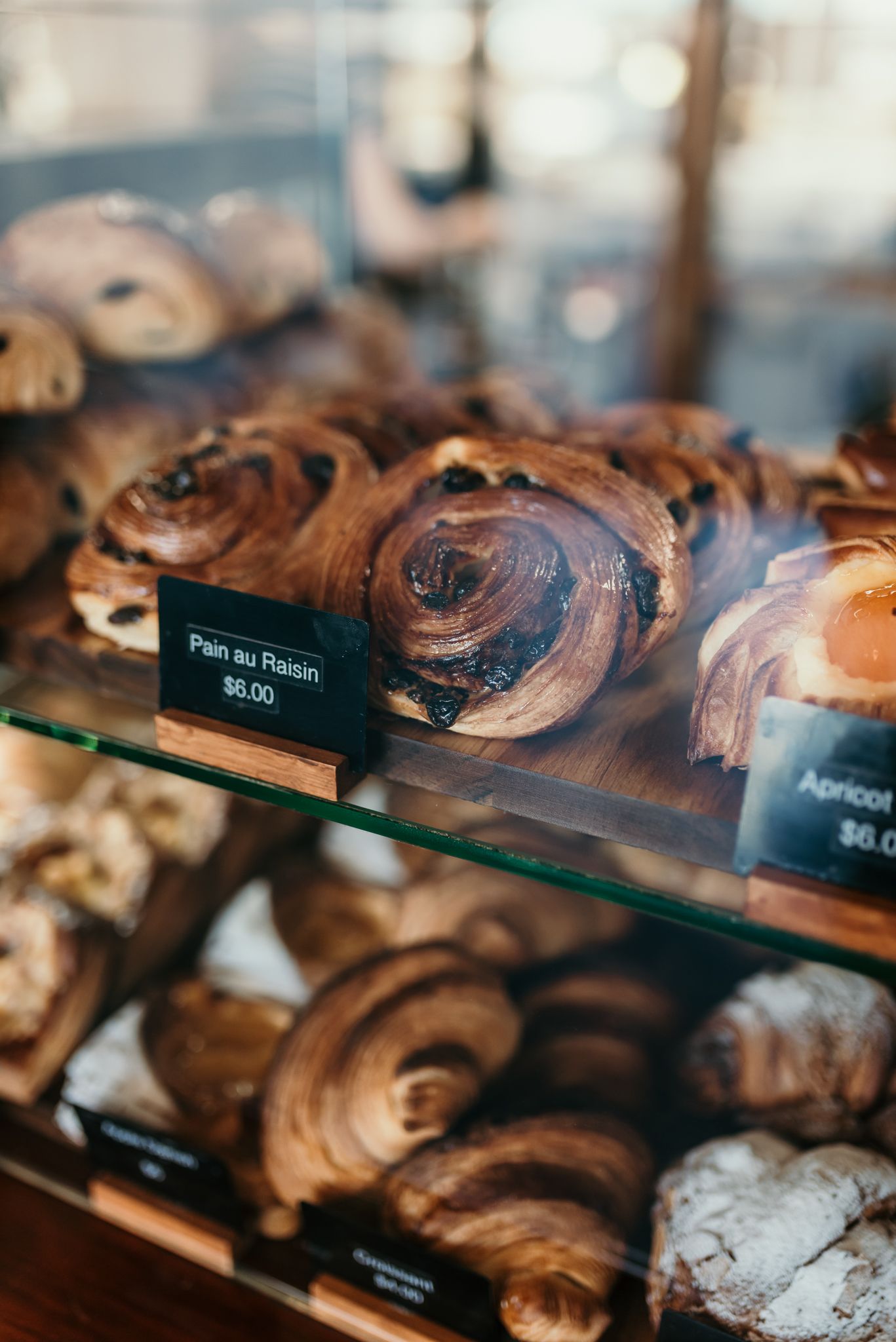 Fresh pain au chocolat pastries displayed behind glass at a bakery with price tag of $6.00.