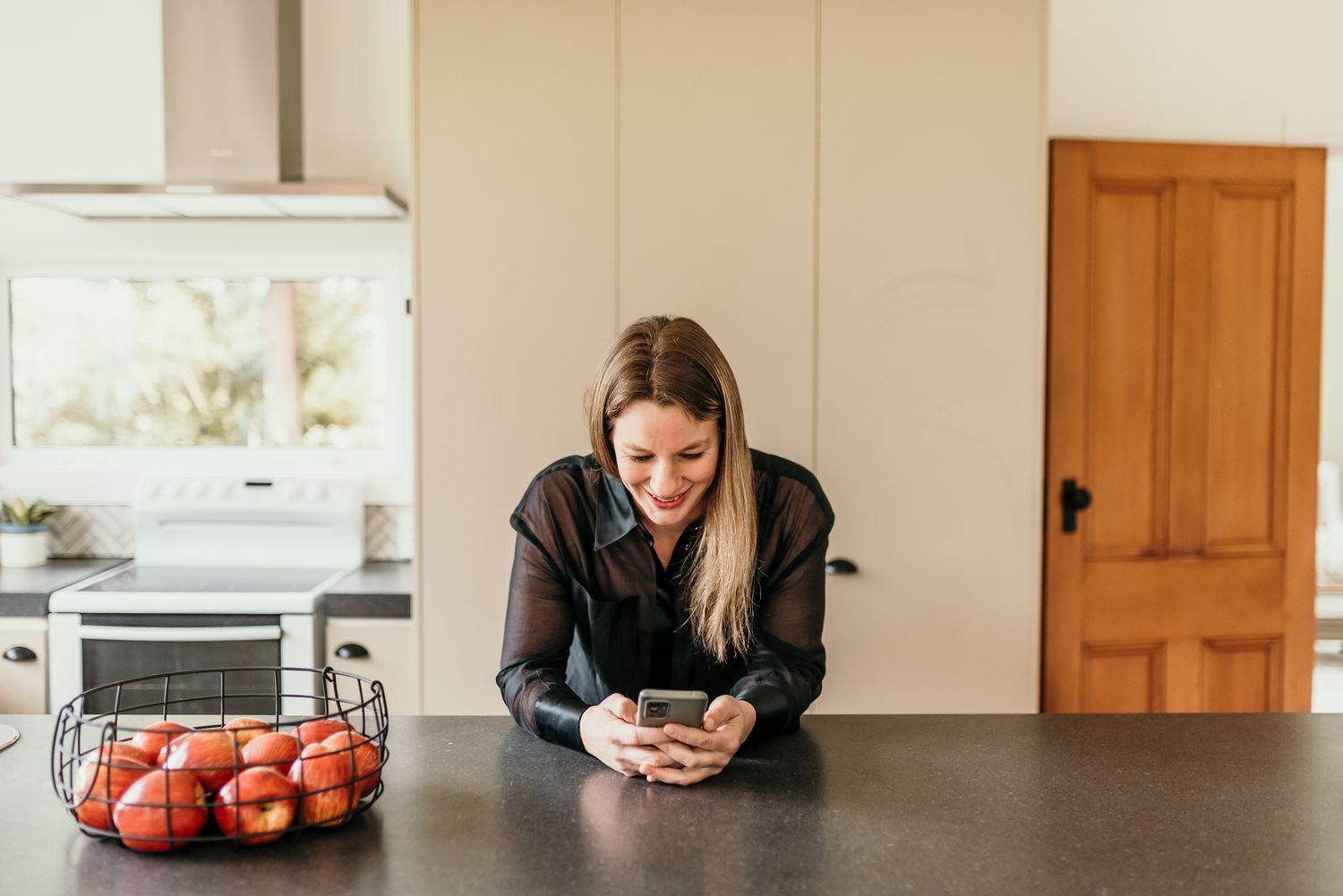 Person checking phone while leaning on kitchen counter with red tomatoes in basket.