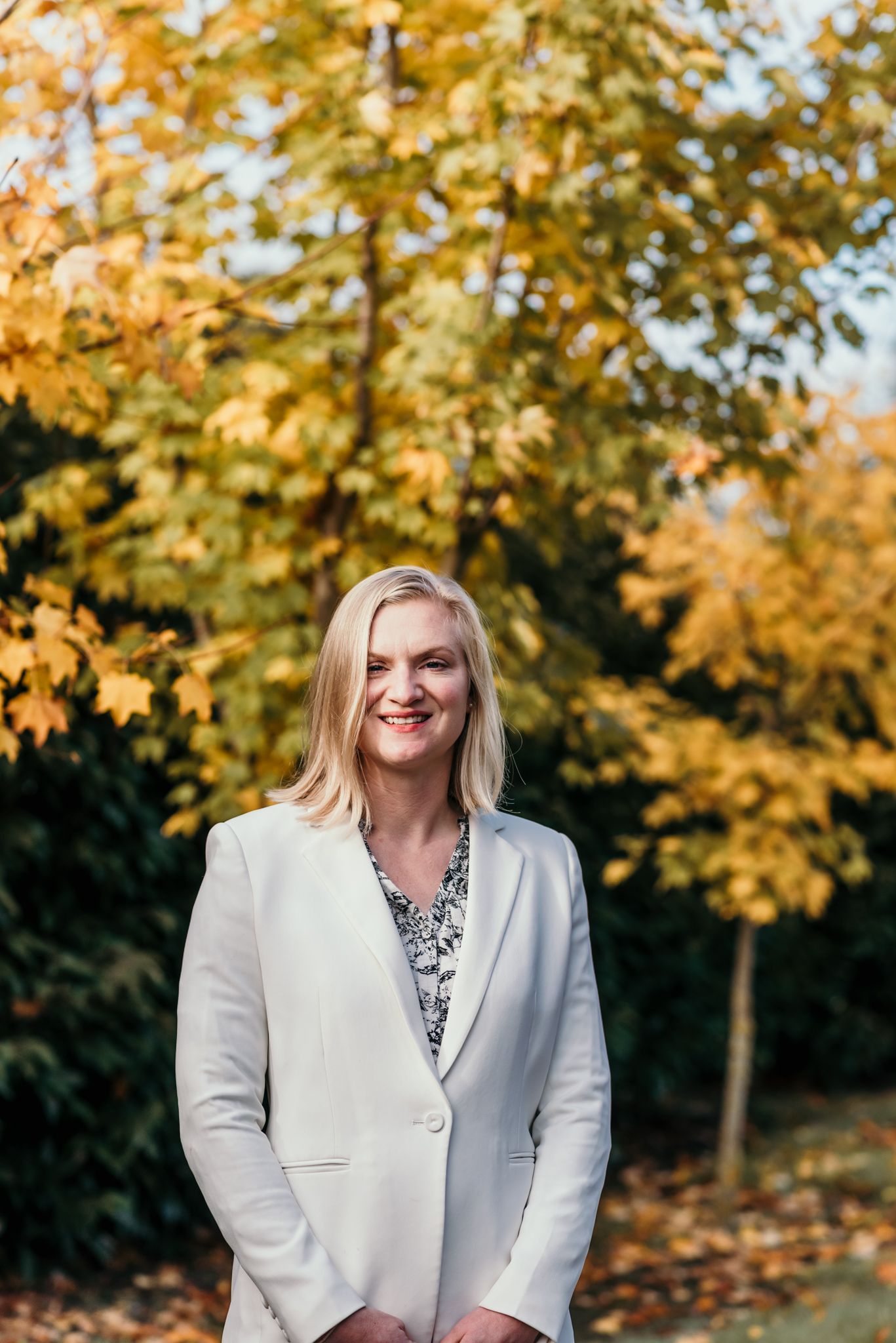 Professional headshot outdoors with autumn foliage in background.