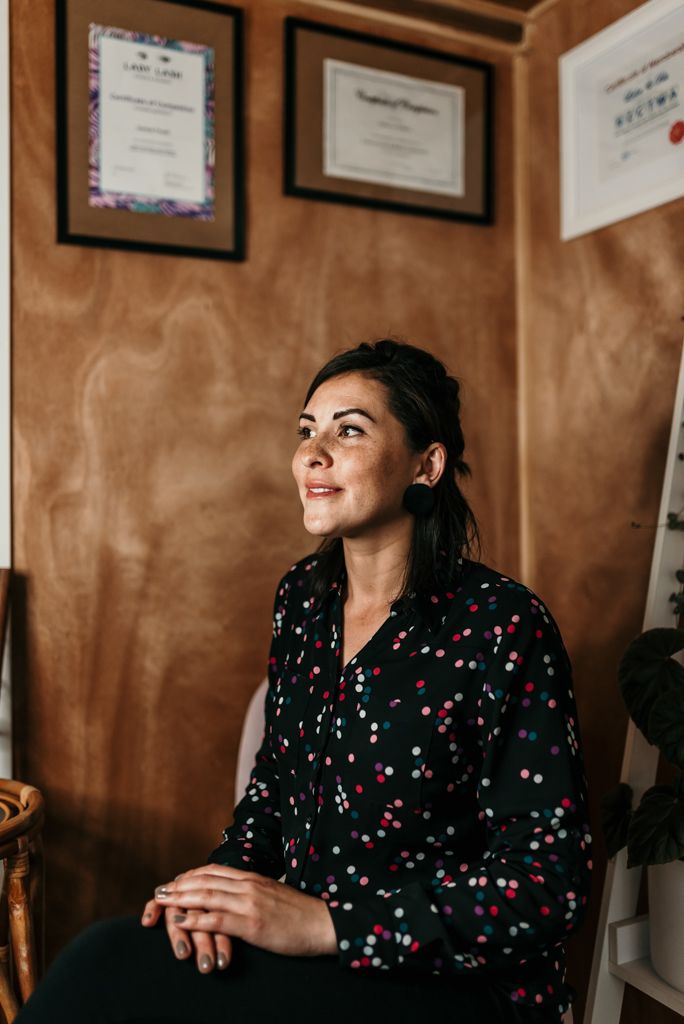 Person in polka dot blouse sitting below framed certificates on brown walls.