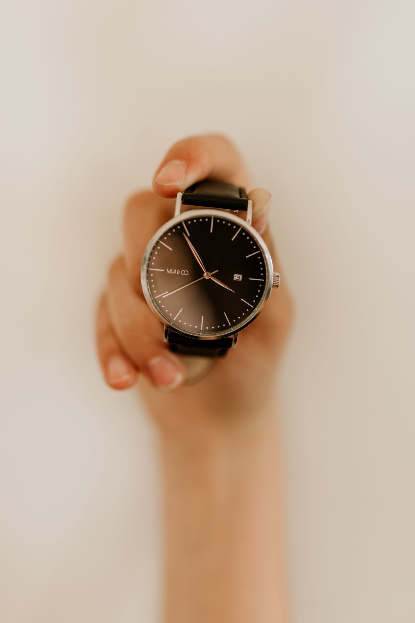 Hand holding up a minimalist black leather watch with silver case against white background.