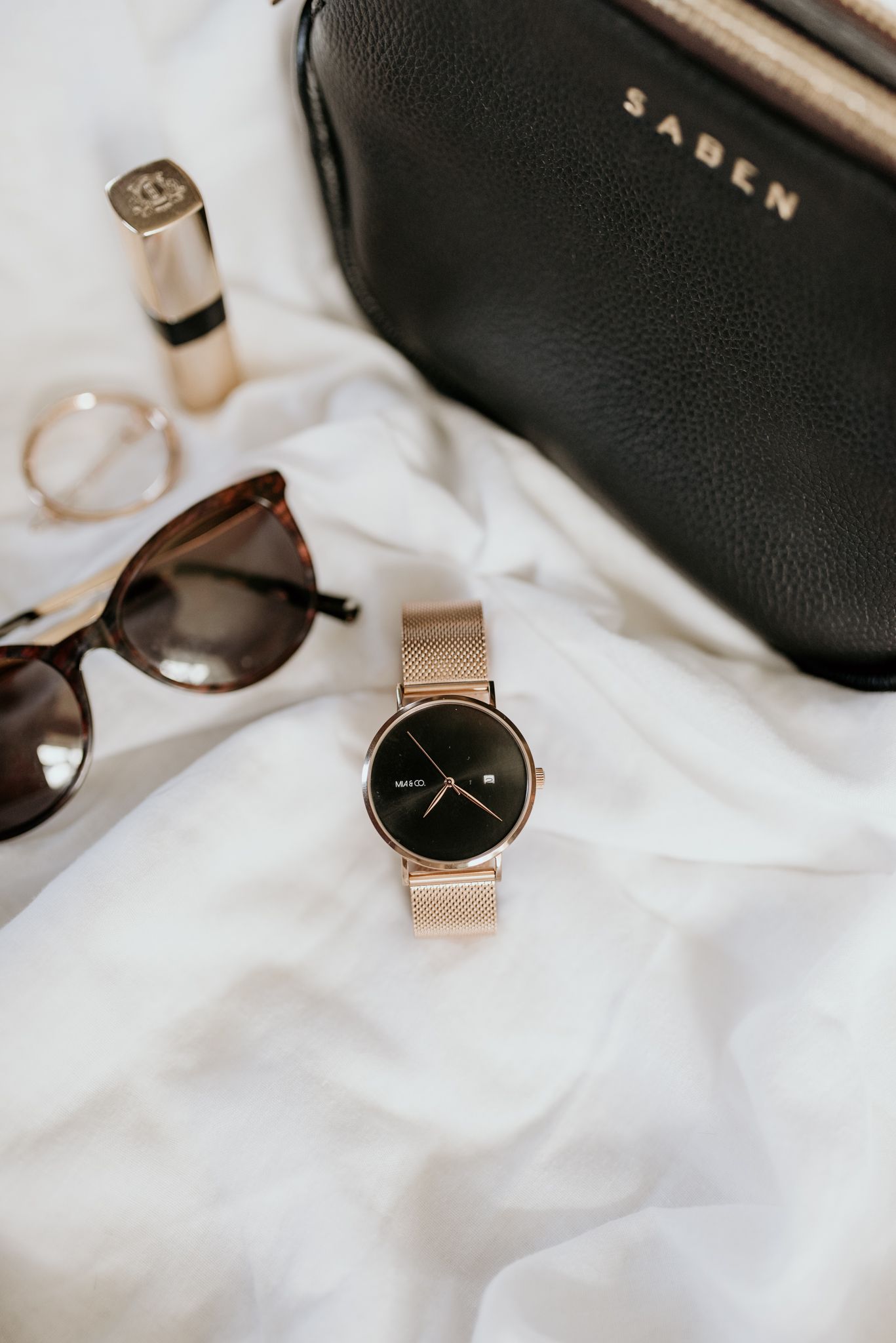 Styled flatlay of accessories including gold watch, sunglasses and black leather bag on white fabric.