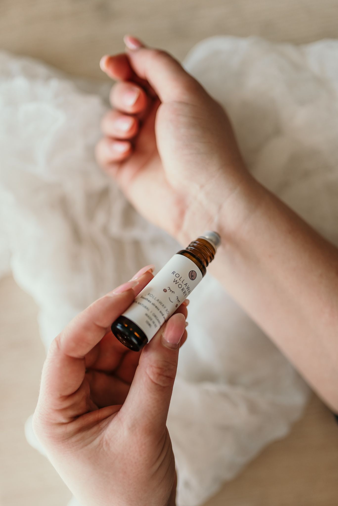 Hands holding a small glass bottle of essential oil or serum against white bedding.