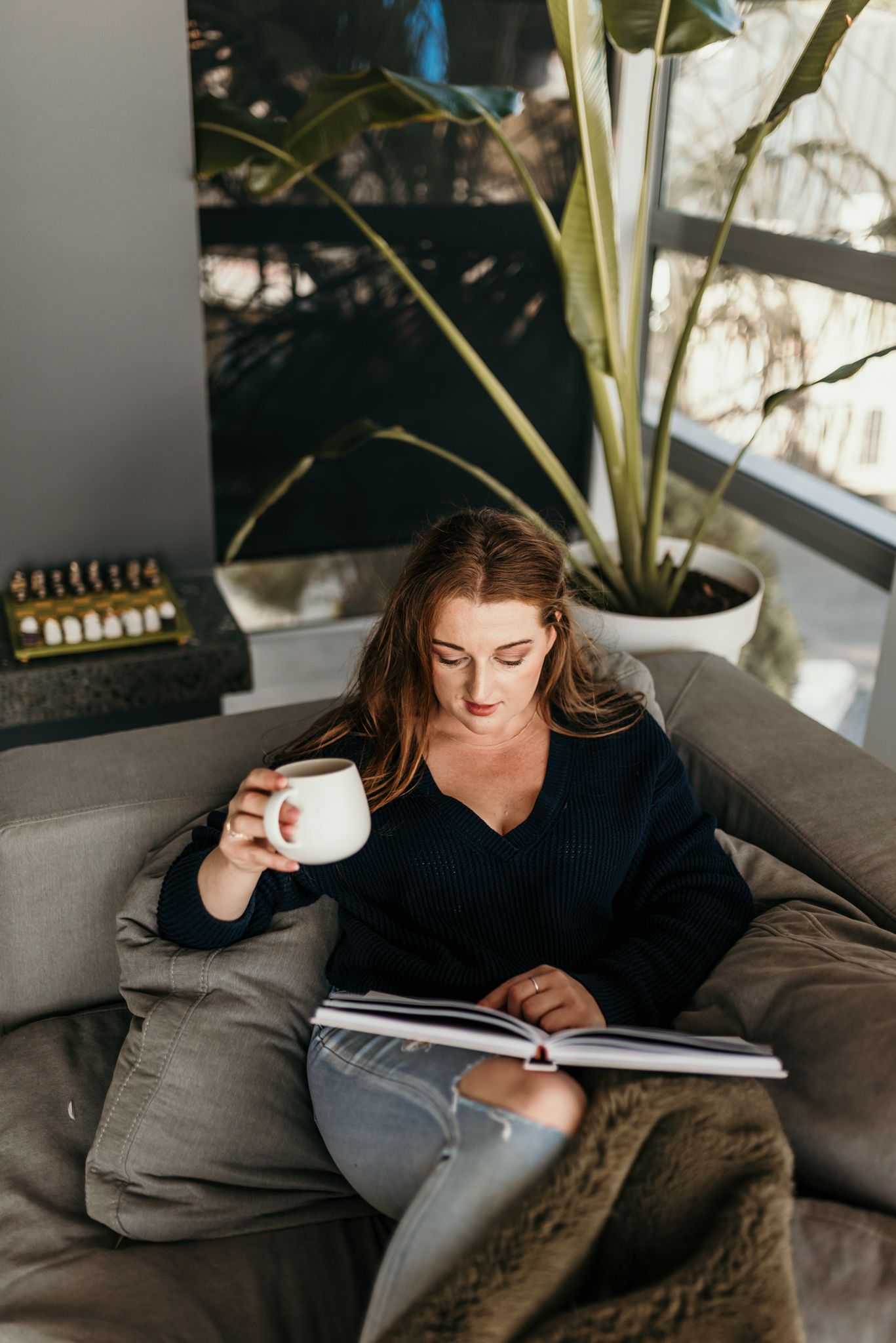Person relaxing on grey couch with coffee cup and book near indoor plant.