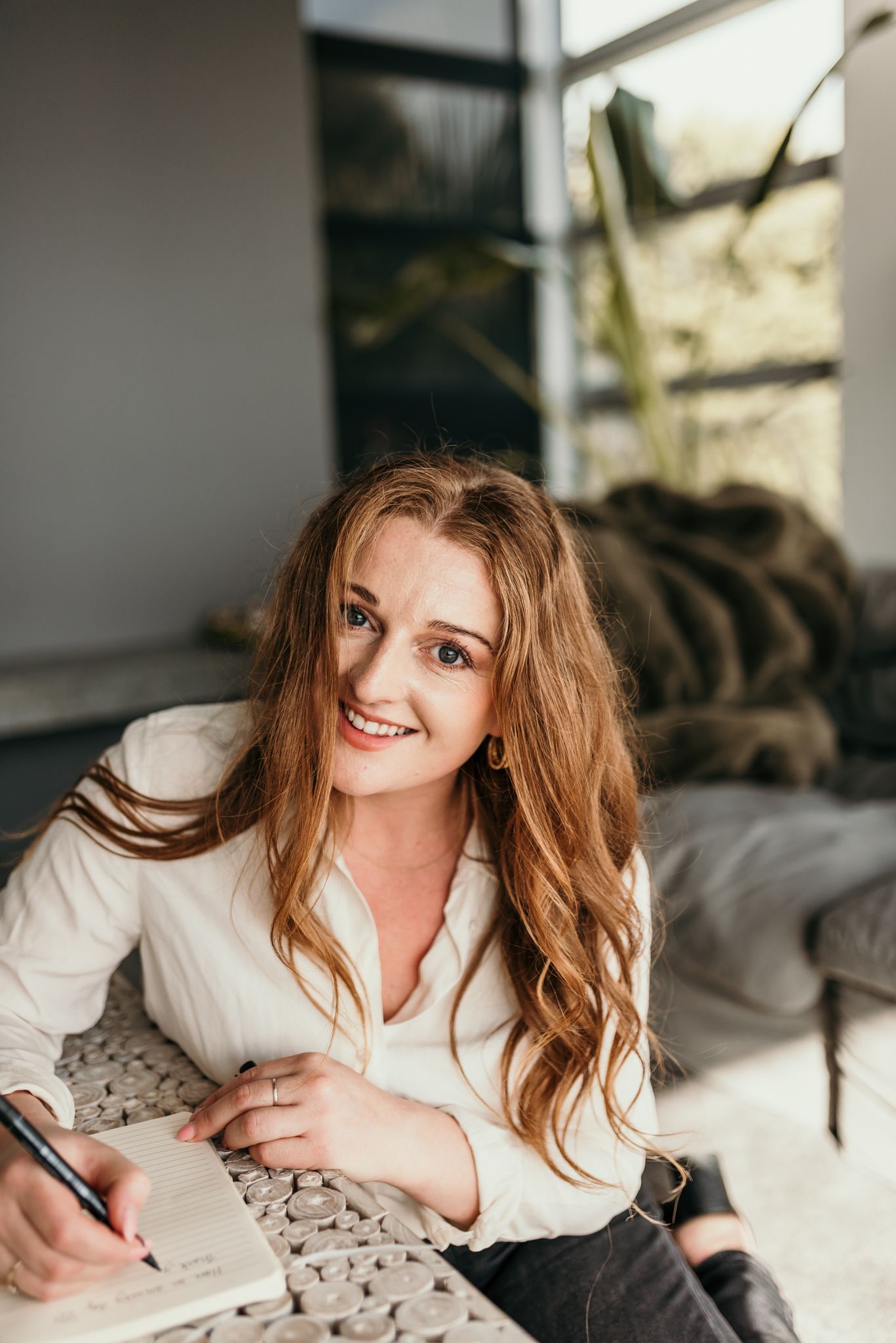 A person in a white shirt writing in a notebook while relaxing on a couch by a window with natural lighting.