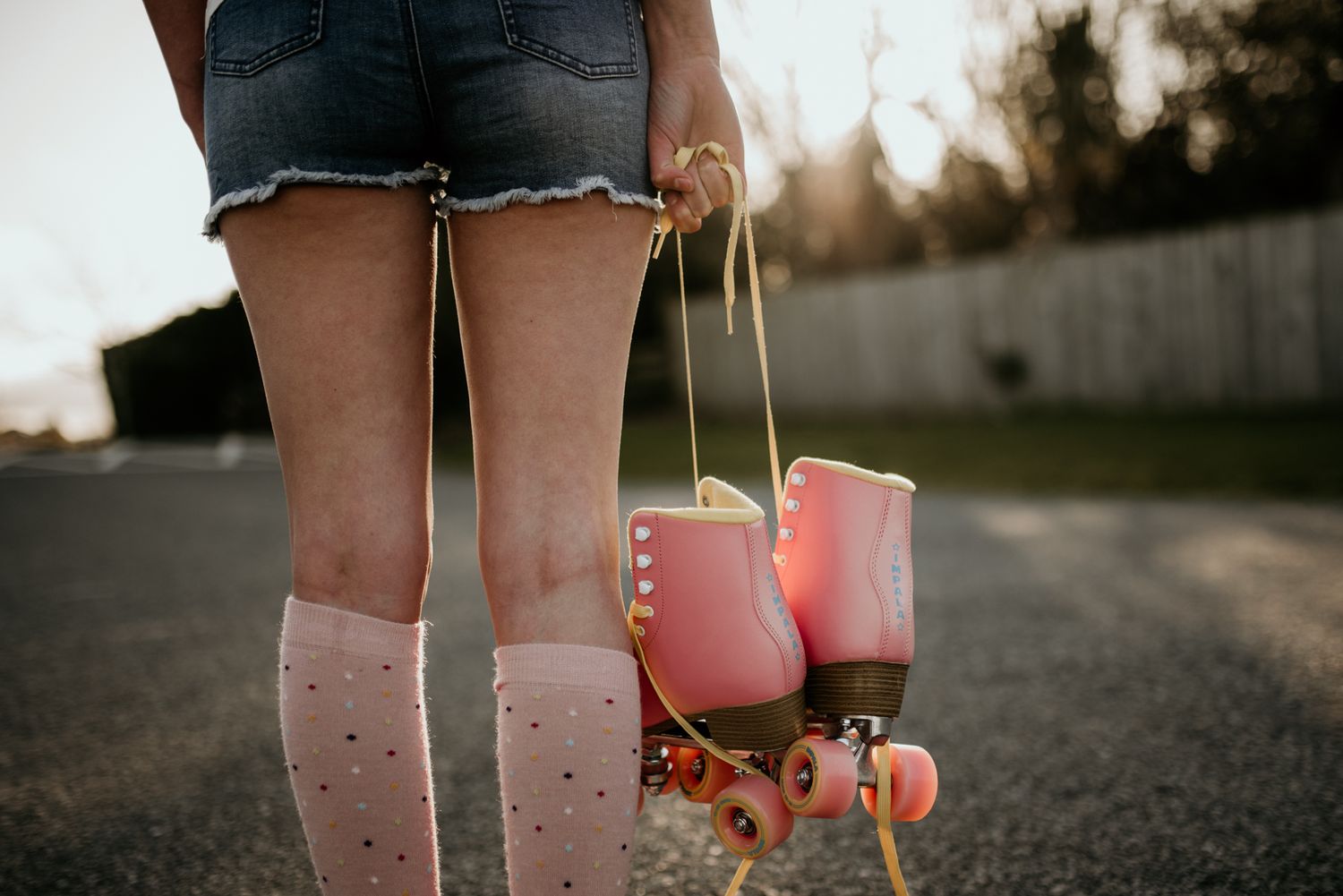 Pink roller skates hang by their laces against a sunset backdrop with polka dot socks visible.